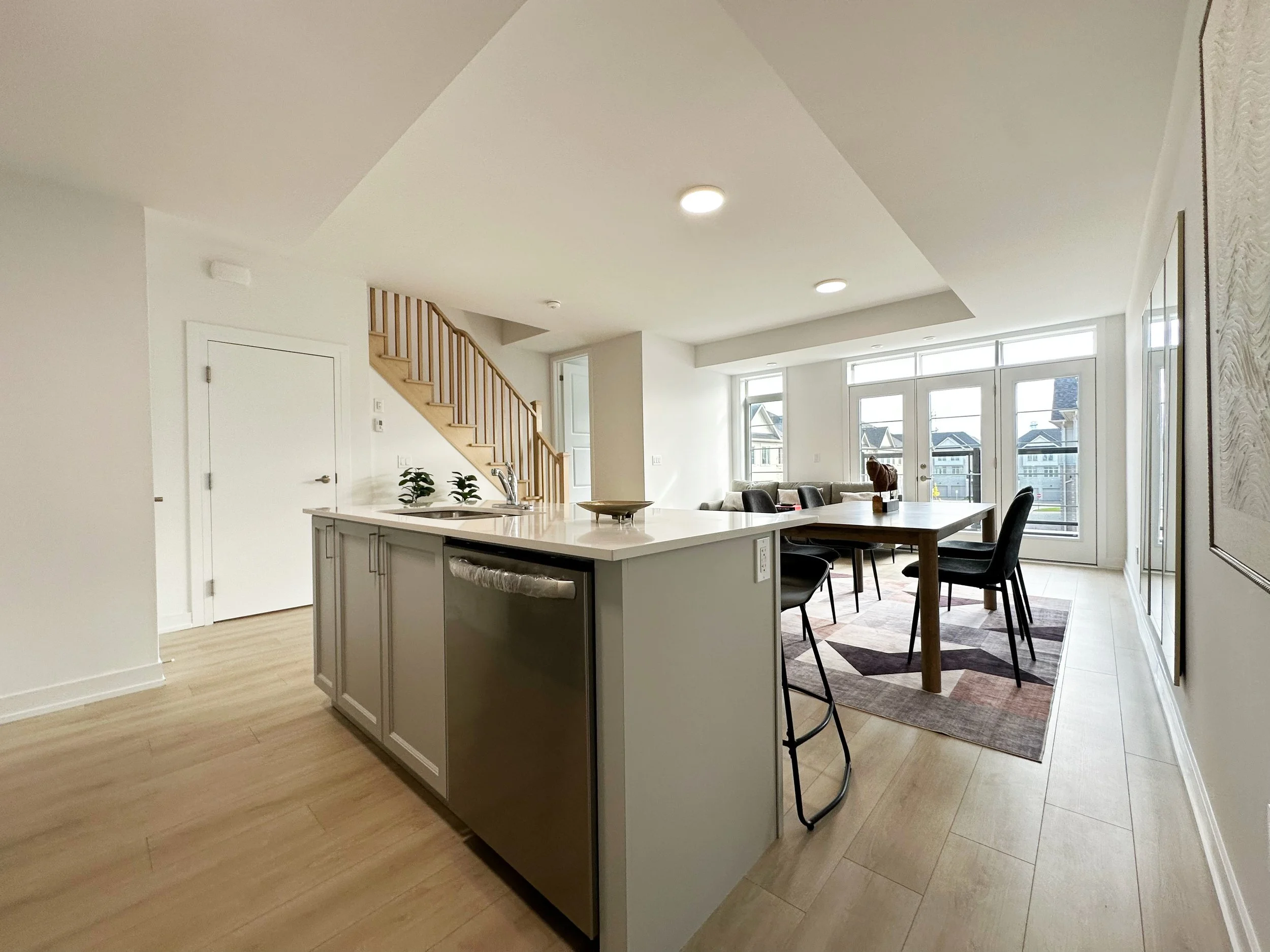 Modern open-concept kitchen with white cabinetry, a large island with a sink, dining area with black chairs, and a living room with large windows and a view of neighboring houses.