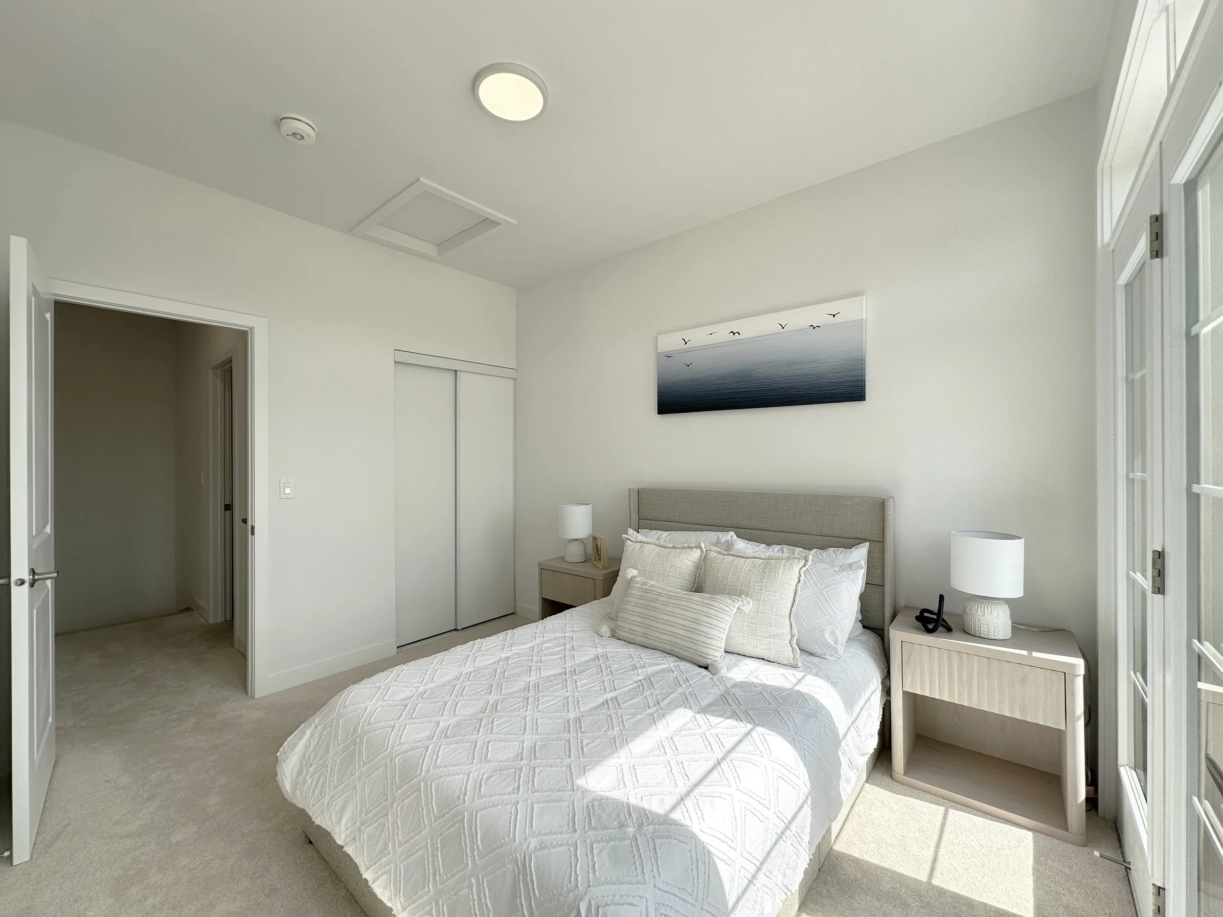 A bedroom with a white bed, a nightstand with a lamp and decorative items, and a wall art piece of birds over water. Sunlight enters through a window, casting shadows.