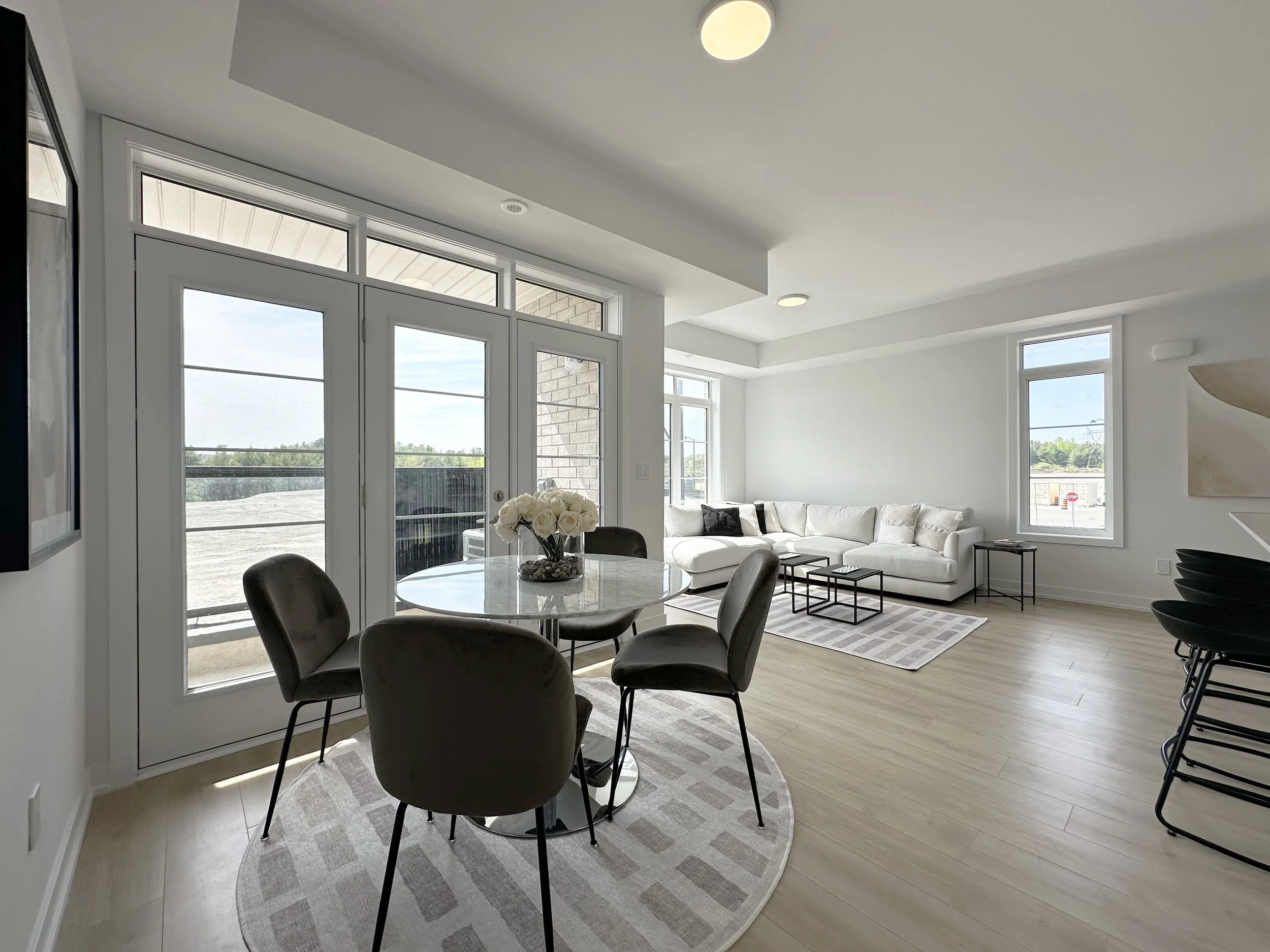 A modern living room and dining area with large windows and glass doors, white walls, a white sofa with black pillows, a round dining table with four chairs, and a flower vase with white roses.