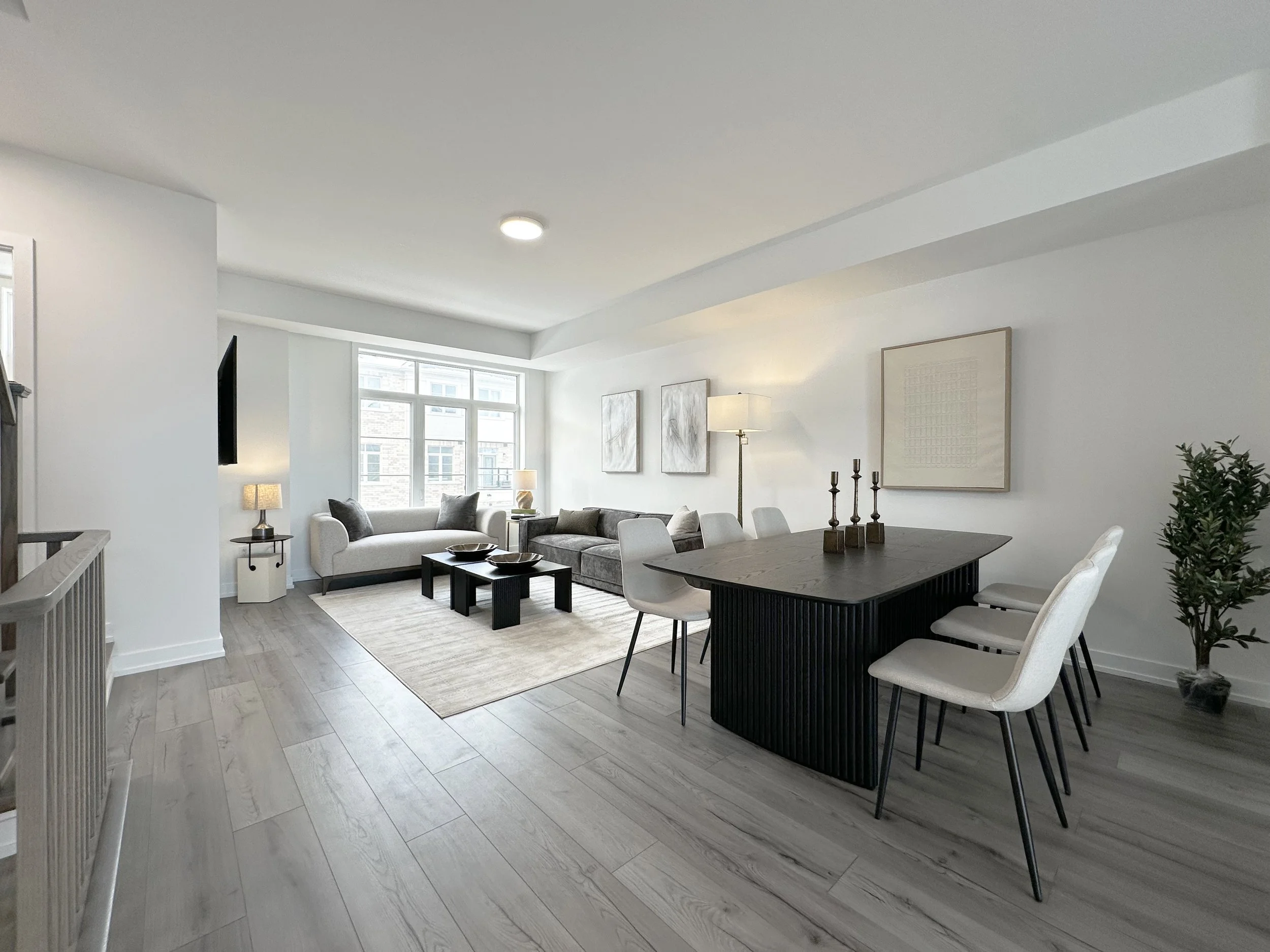 A spacious living and dining area with white walls, large windows, and contemporary furniture including a sofa, chairs, and a dining table with chairs. There are decorative elements like framed artwork, lamps, and candles.