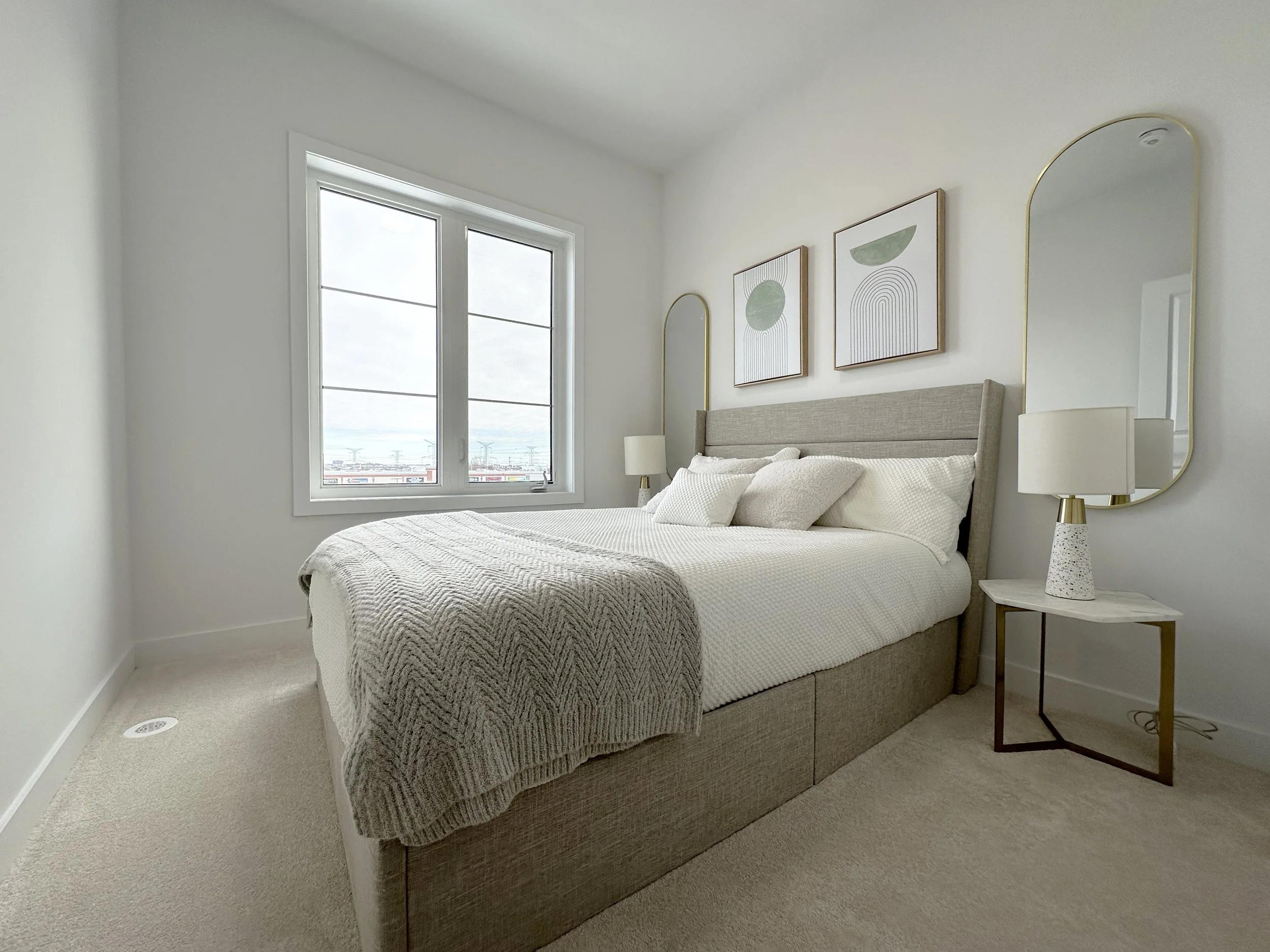 A cozy bedroom with a beige upholstered bed, white bedding, and decorative pillows. Two tall, elegant table lamps with white shades sit on nightstands beside the bed. Two framed abstract art pieces hang above the headboard, and two tall, gold-framed 