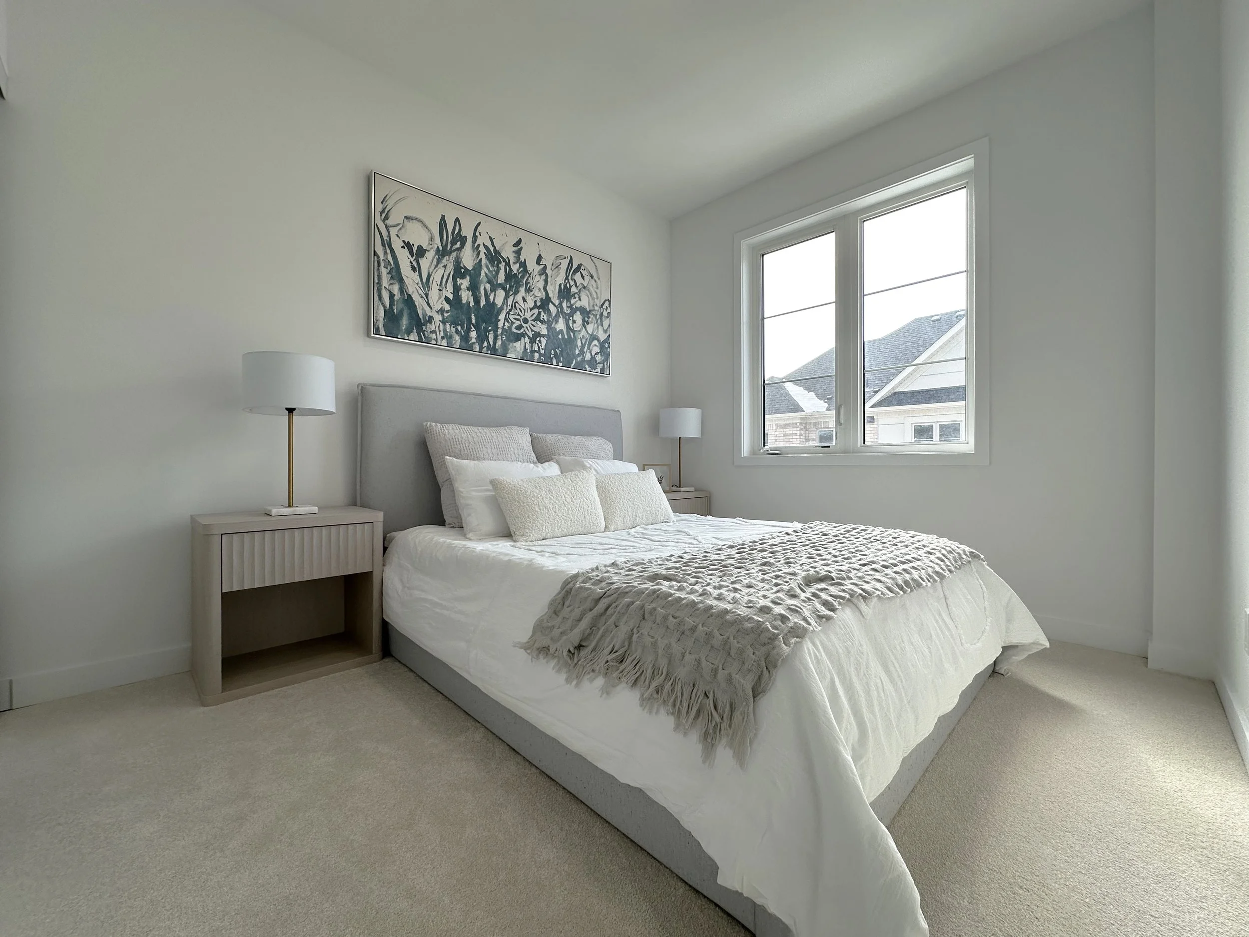 A bedroom with a white bed, beige throw blanket, and multiple pillows. Beside the bed are matching white nightstands with white lamps. There is a large window showing rooftops outside and a framed abstract black and white painting on the wall.
