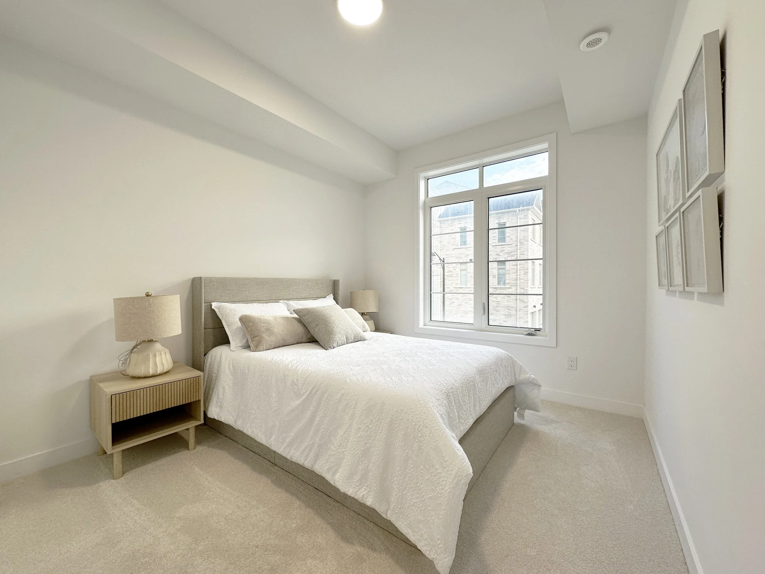 A bedroom with a bed, two beige lamps, a beige headboard, and a large window showing a building outside.