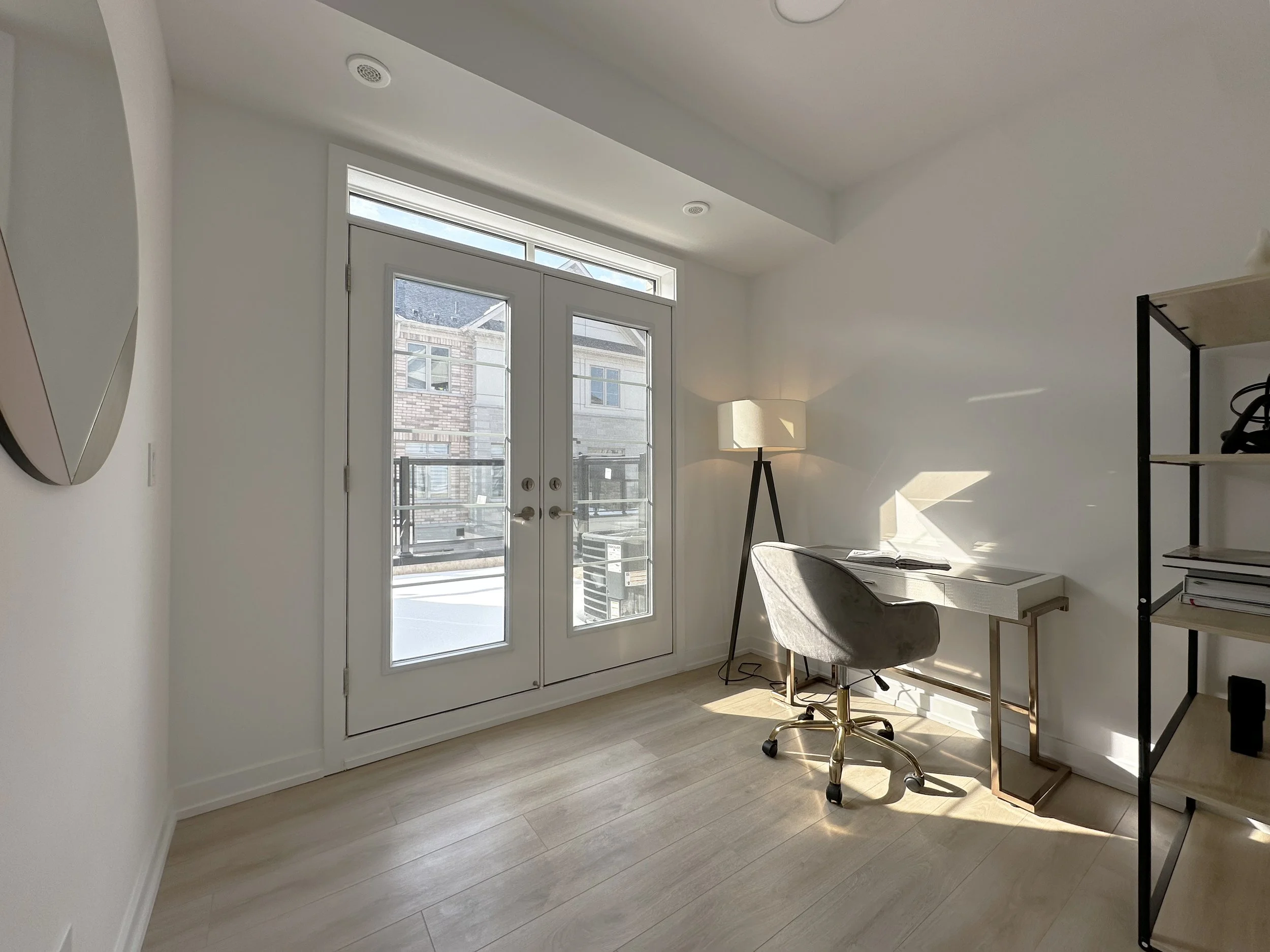 Bright room with large glass doors leading outside, a lamp, a chair, and a shelving unit, with sunlight casting shadows on the beige wooden floor.