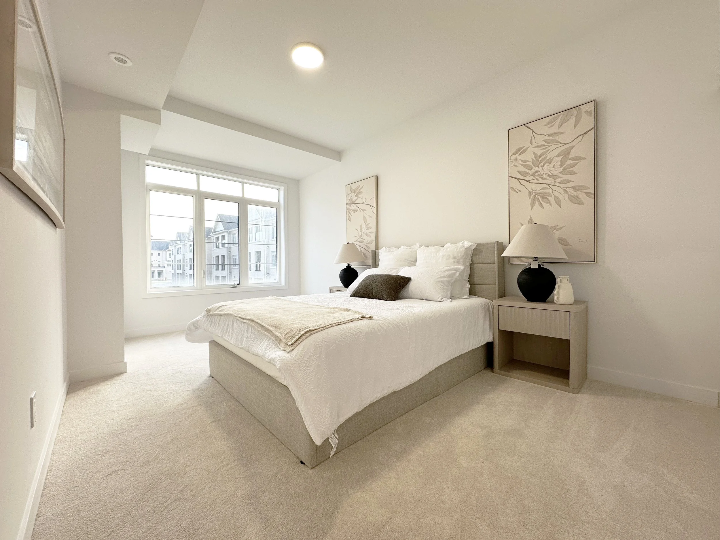 A bedroom with a bed, two nightstands with lamps, and framed artwork on the walls. There are large windows showing neighboring buildings outside.