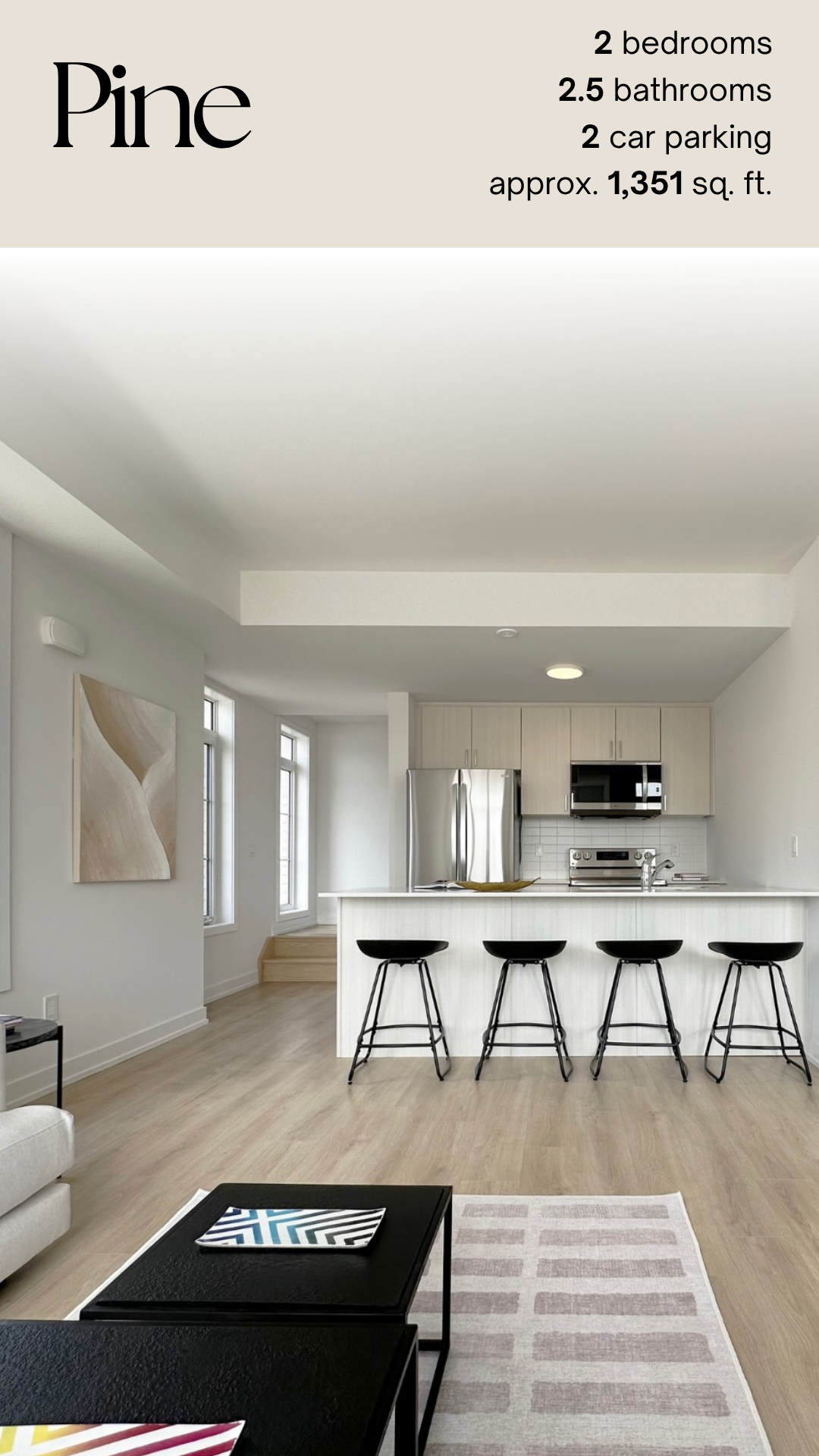 Modern kitchen with white cabinets, stainless steel appliances, a white island with black chairs, and a light wood floor. There is artwork on the wall and windows letting in natural light.