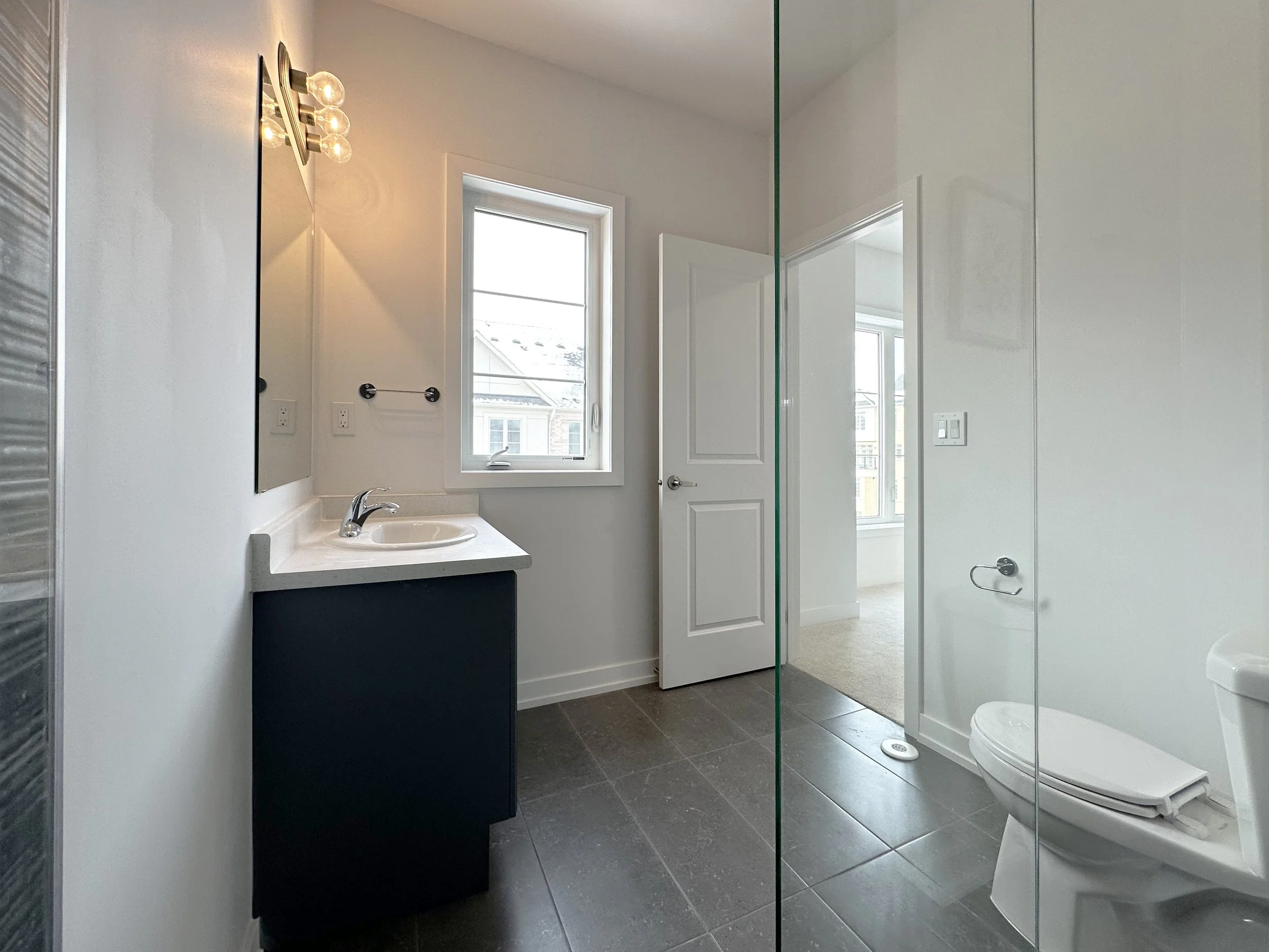 Modern bathroom with a white toilet, a black vanity with a white countertop and sink, a wall-mounted mirror, a window for natural light, and a glass shower door.