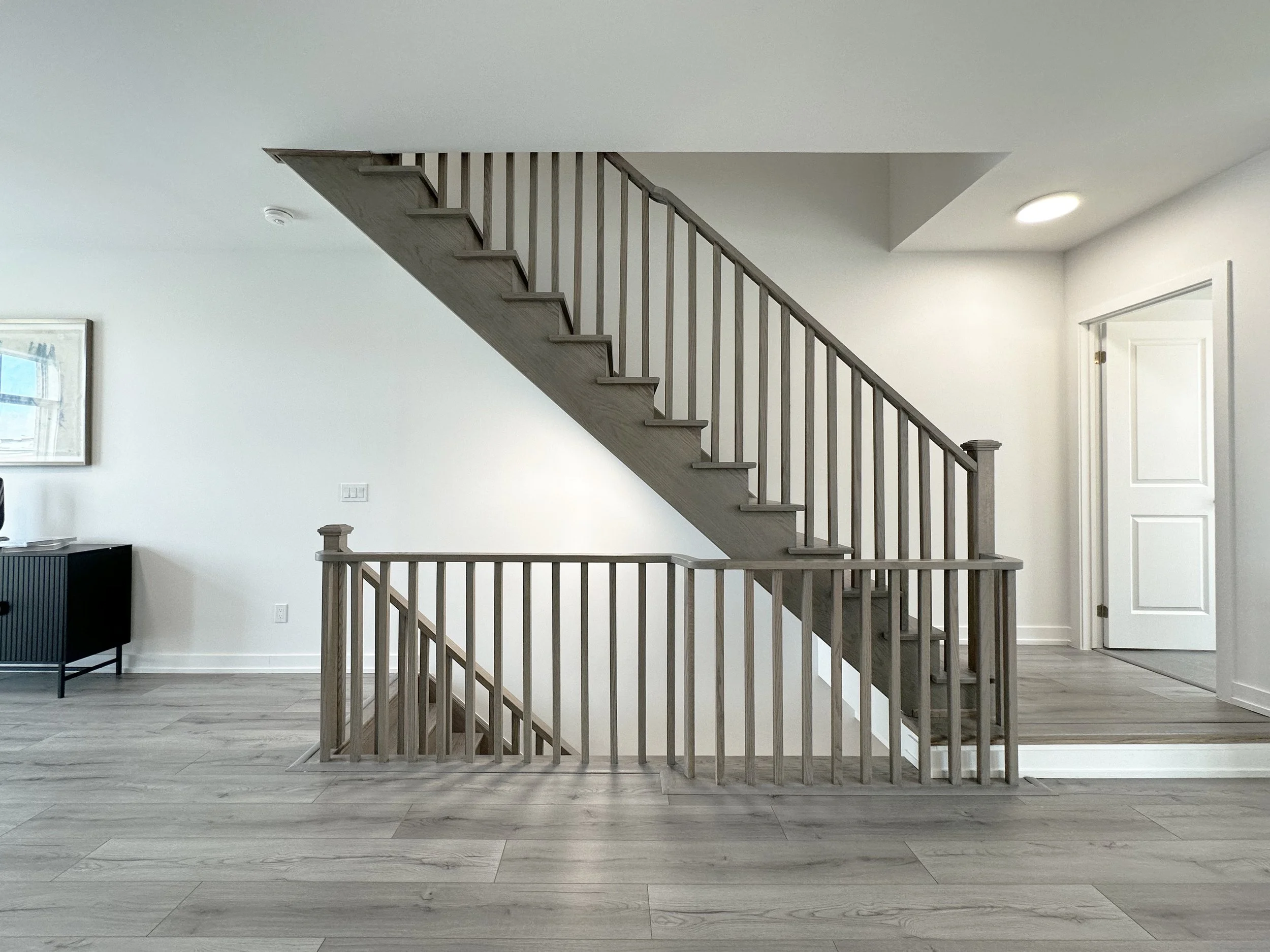 Interior view of a modern house showing a staircase with wooden railings and steps, white walls, a door on the right, a window on the left, a framed picture on the wall, a ceiling light, and a smoked glass side table.