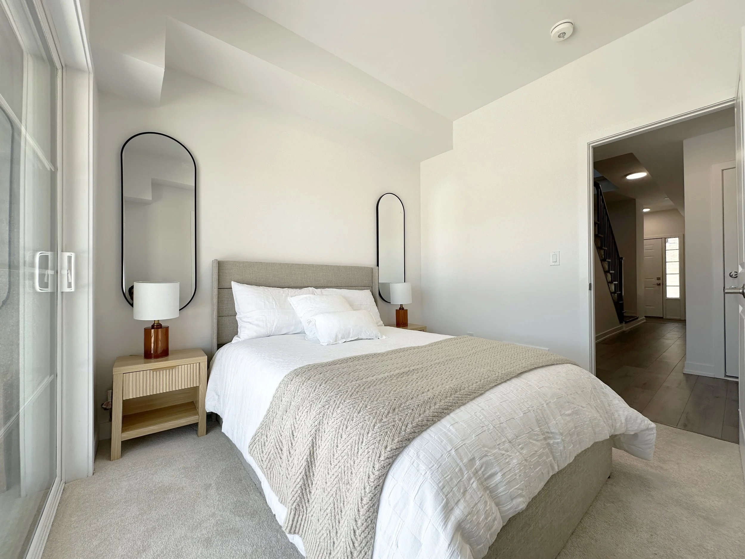 A bedroom with a bed, bedside tables with lamps, and mirrors on the wall. There is an open doorway leading to a hallway with stairs.