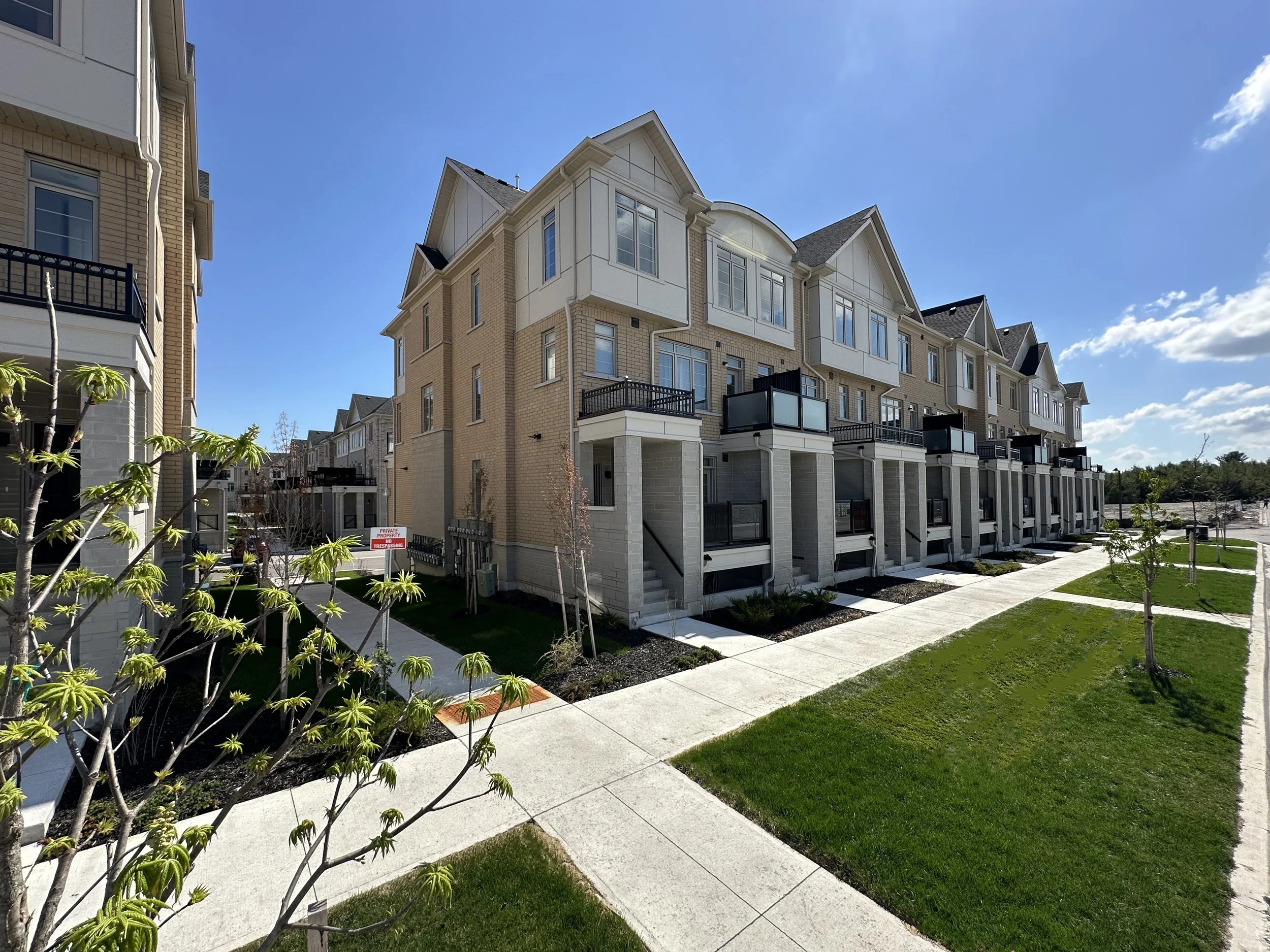 Newly built multi-story residential buildings with balconies, surrounded by green lawns, sidewalks, and small trees under a bright blue sky with some clouds.