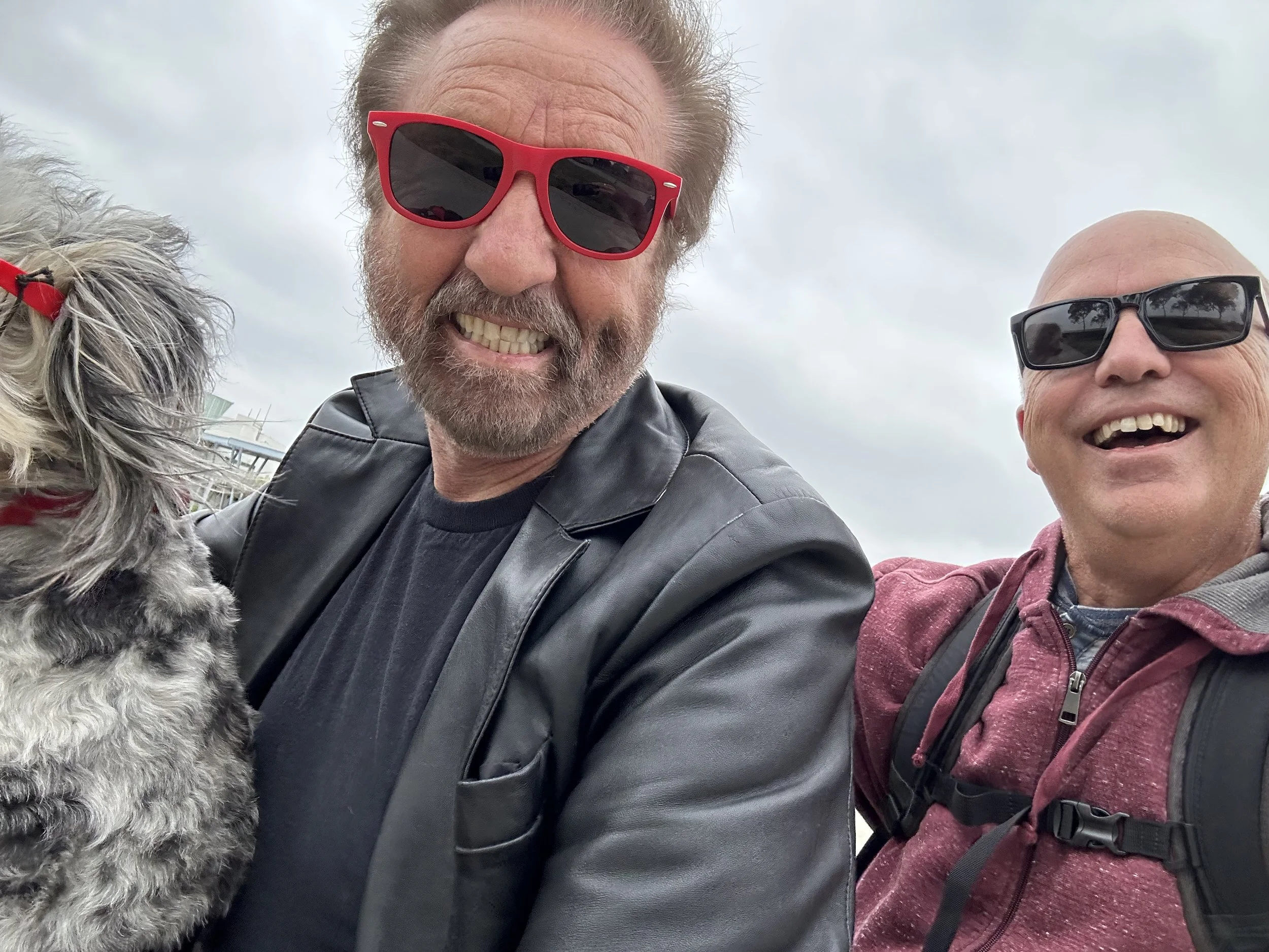 Three people and a dog taking a selfie outdoors on a cloudy day. The person in the middle is a man with a beard wearing red sunglasses and a black leather jacket. To his right is a man with black sunglasses and a maroon jacket. The dog, with curly gray fur, is on the left side of the image, partially visible.