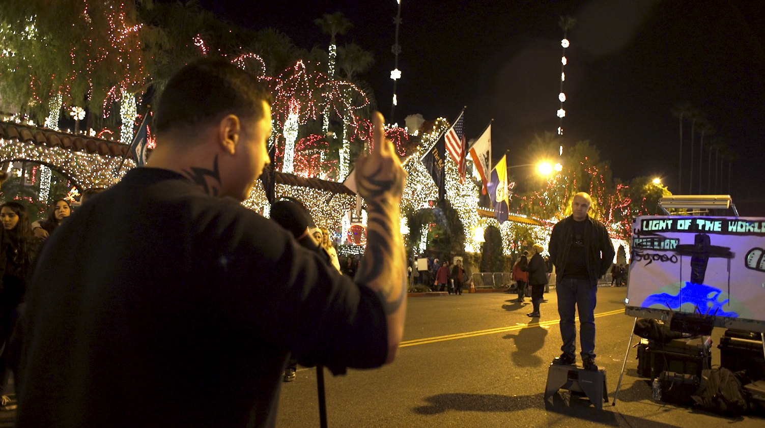 A man with tattoos on his neck and arms is holding a microphone and singing or speaking into it at a nighttime outdoor event with colorful holiday lights and trees decorated in white and red lights in the background. Several other people are gathered around, and there are flags and a digital display showing artwork related to the theme of light.