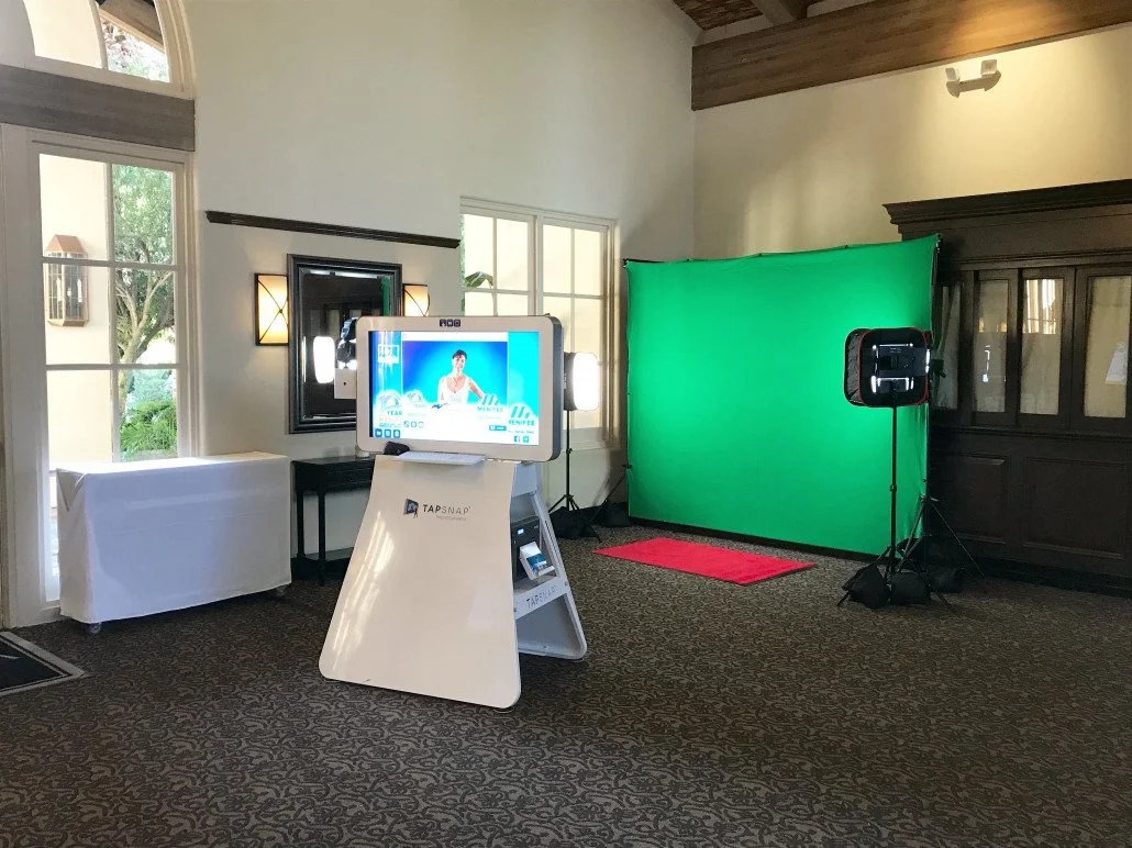 Event photo booth setup with green screen backdrop, studio lights, touchscreen kiosk, and red carpet in an elegant indoor venue