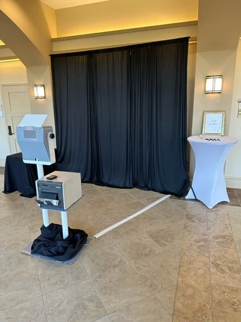 Event photo booth with printer in front of black curtains, beside a cocktail table and framed sign in a hotel lobby