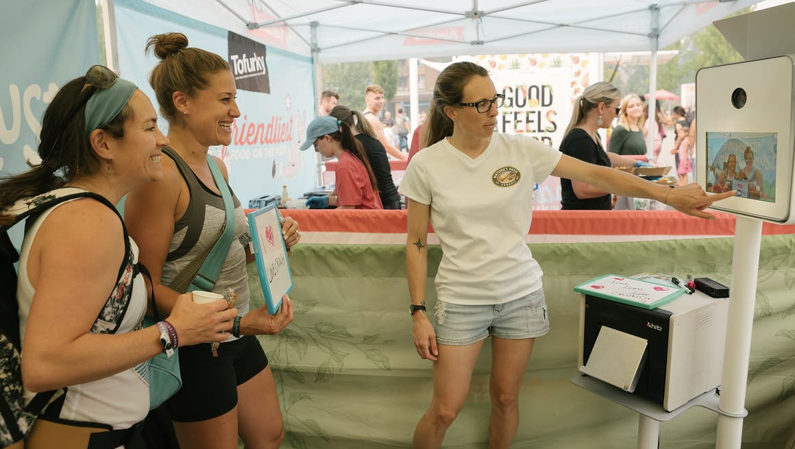 Women visit a Tot Tury’s promotional booth with a staff member demonstrating a touchscreen photo kiosk.