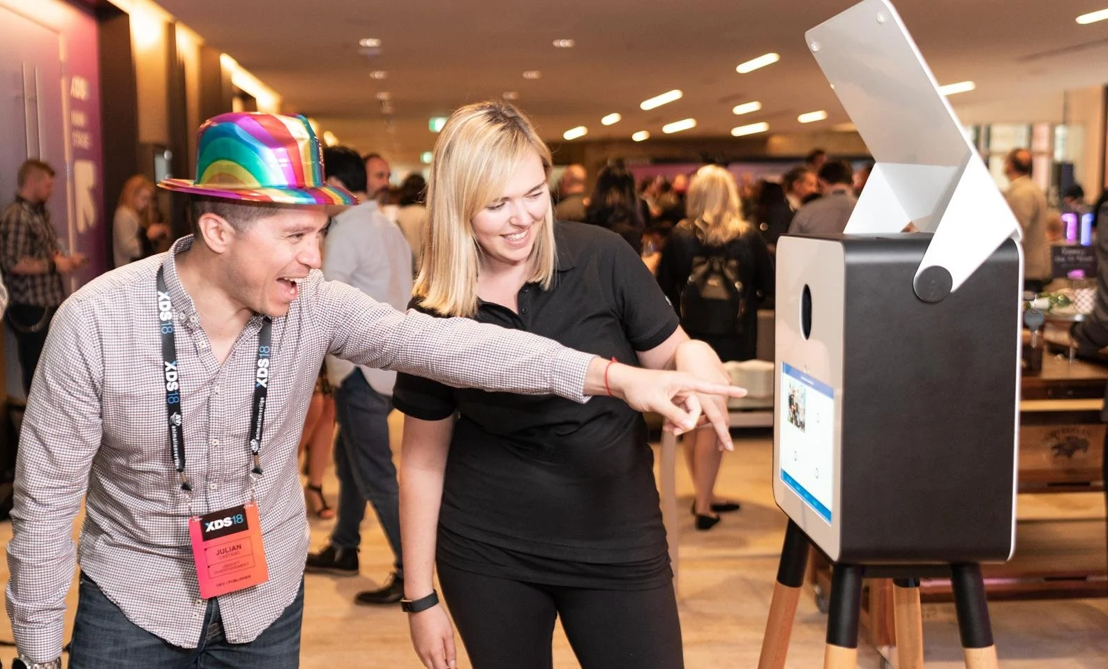 Two people interact with a large touchscreen kiosk at a busy indoor event.