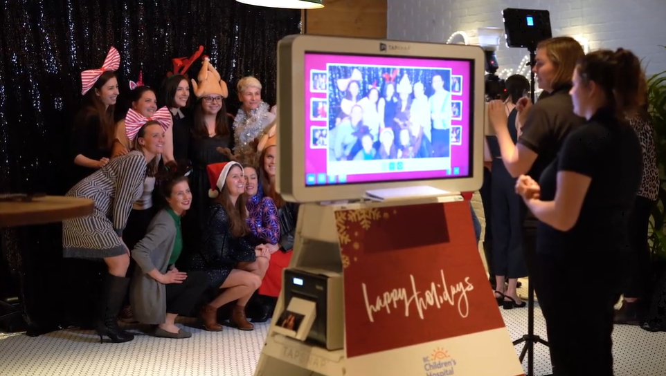 Holiday photo booth capturing a group of women wearing festive accessories beside a Happy Holidays sign.