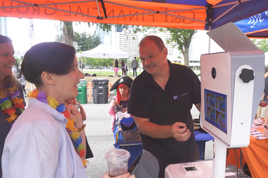 People demonstrating a TapSnap photo booth at an outdoor event under a colorful tent.