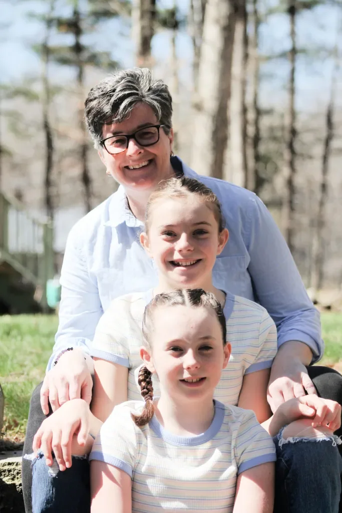 A woman and two young girls sitting outdoors on grass with trees in the background, smiling at the camera.