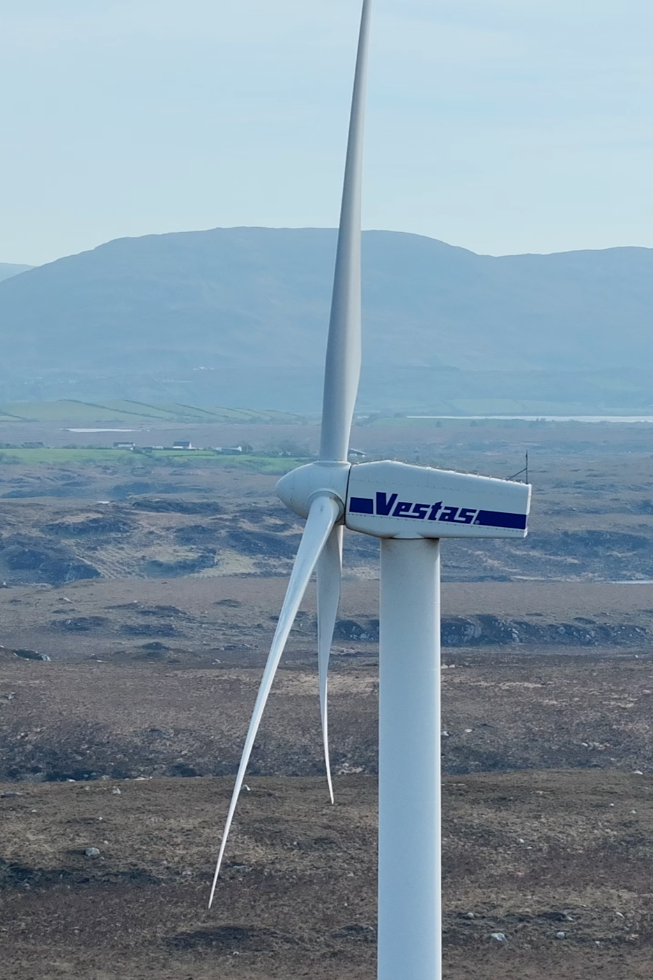 A white wind turbine with the logo 'Vestas' on the side, standing on a barren landscape with hills and mountains in the background.