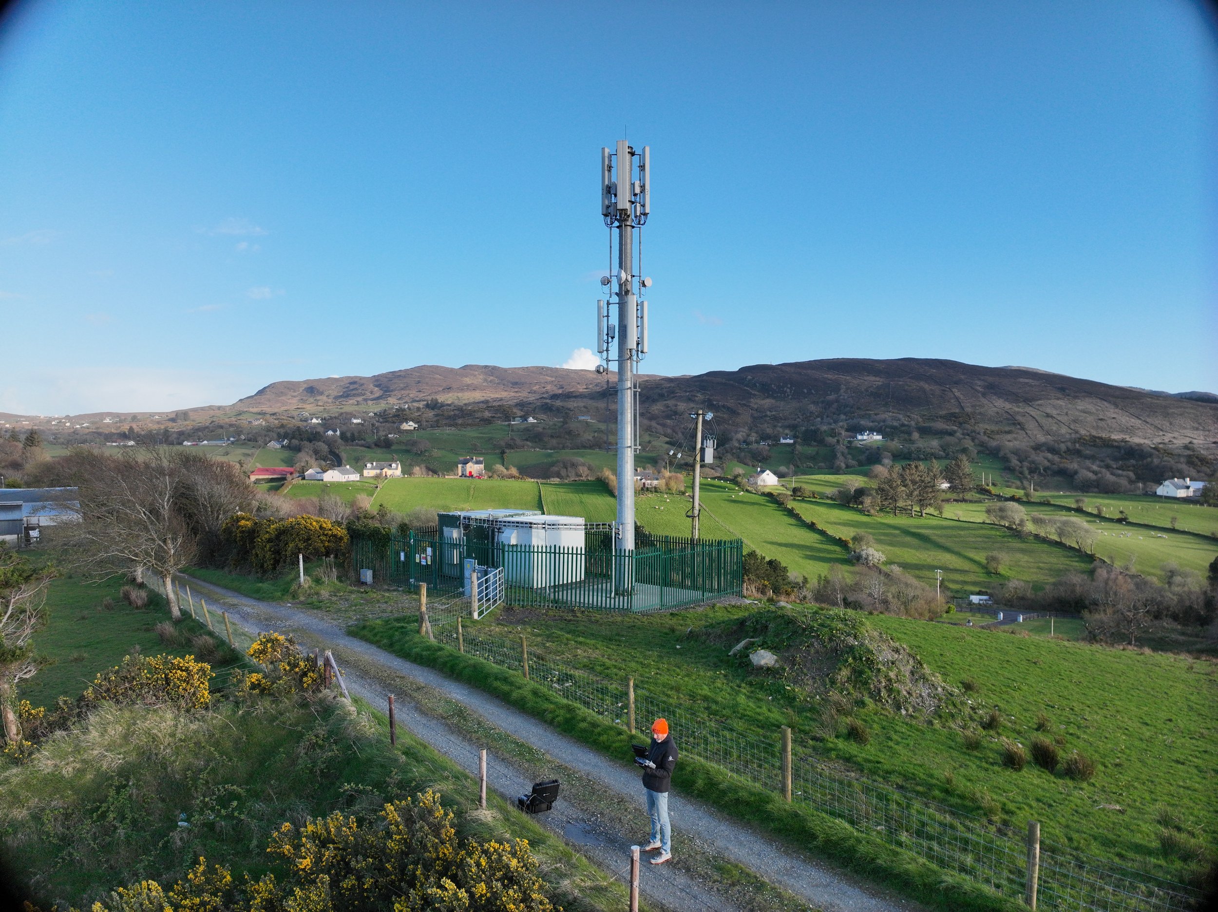 A person wearing an orange safety helmet and safety clothing working outdoors on a grassy hillside, with telecom equipment on a metal tower and a small building nearby, under a clear blue sky with mountains in the background.