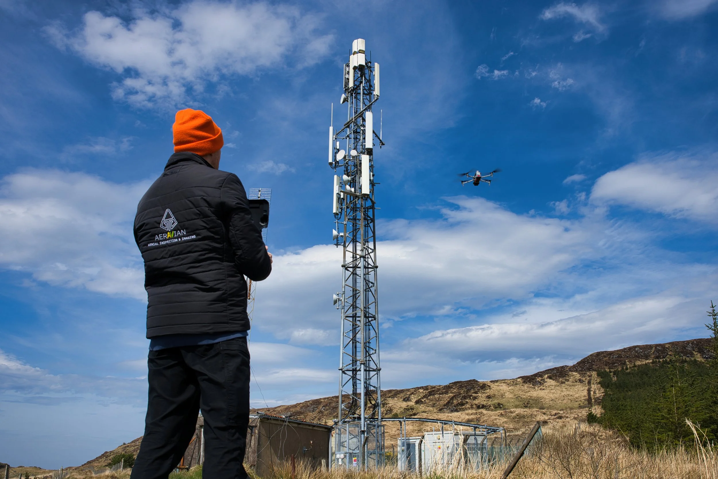 A person in a black jacket and orange beanie operating a drone next to a large cellular tower in a rural area with hills and a partly cloudy sky.