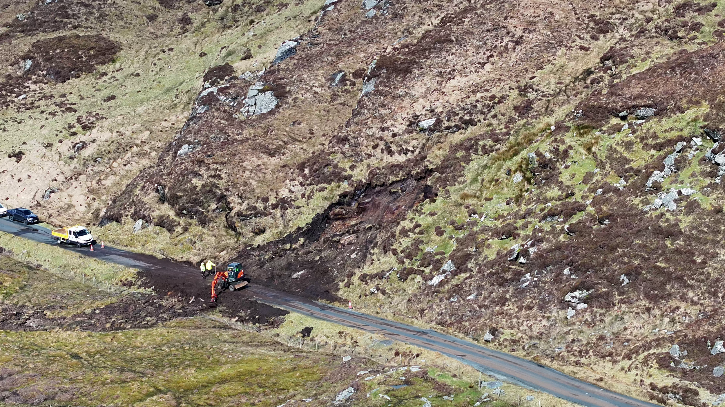 View of a hillside with ongoing road construction; construction vehicles and workers are on the road while the hillside behind shows patches of grass, rocks, and dirt.