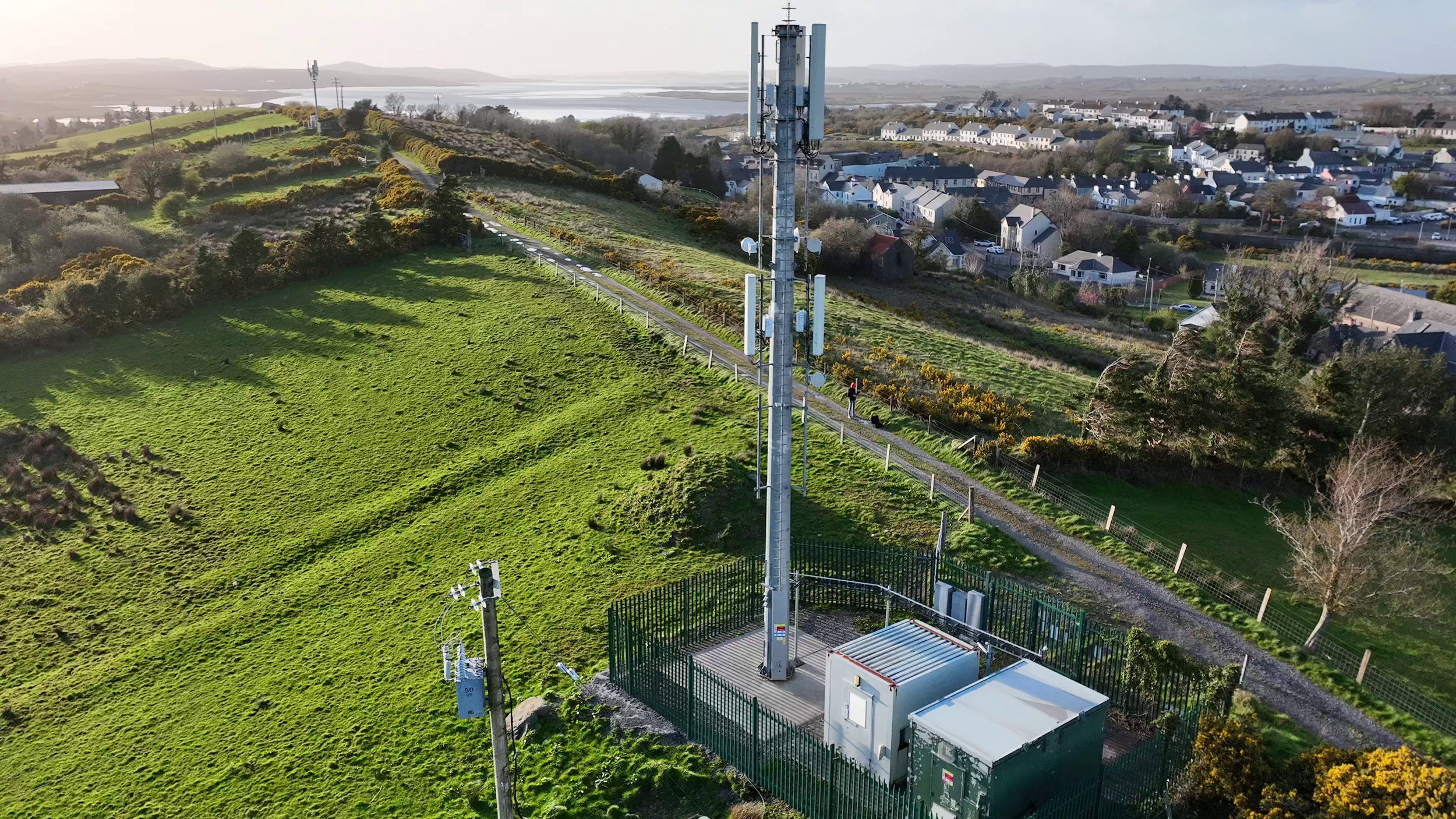 A tall communication antenna tower with various antennas and equipment, enclosed by a secure fence, situated on a grassy hillside near a residential area.