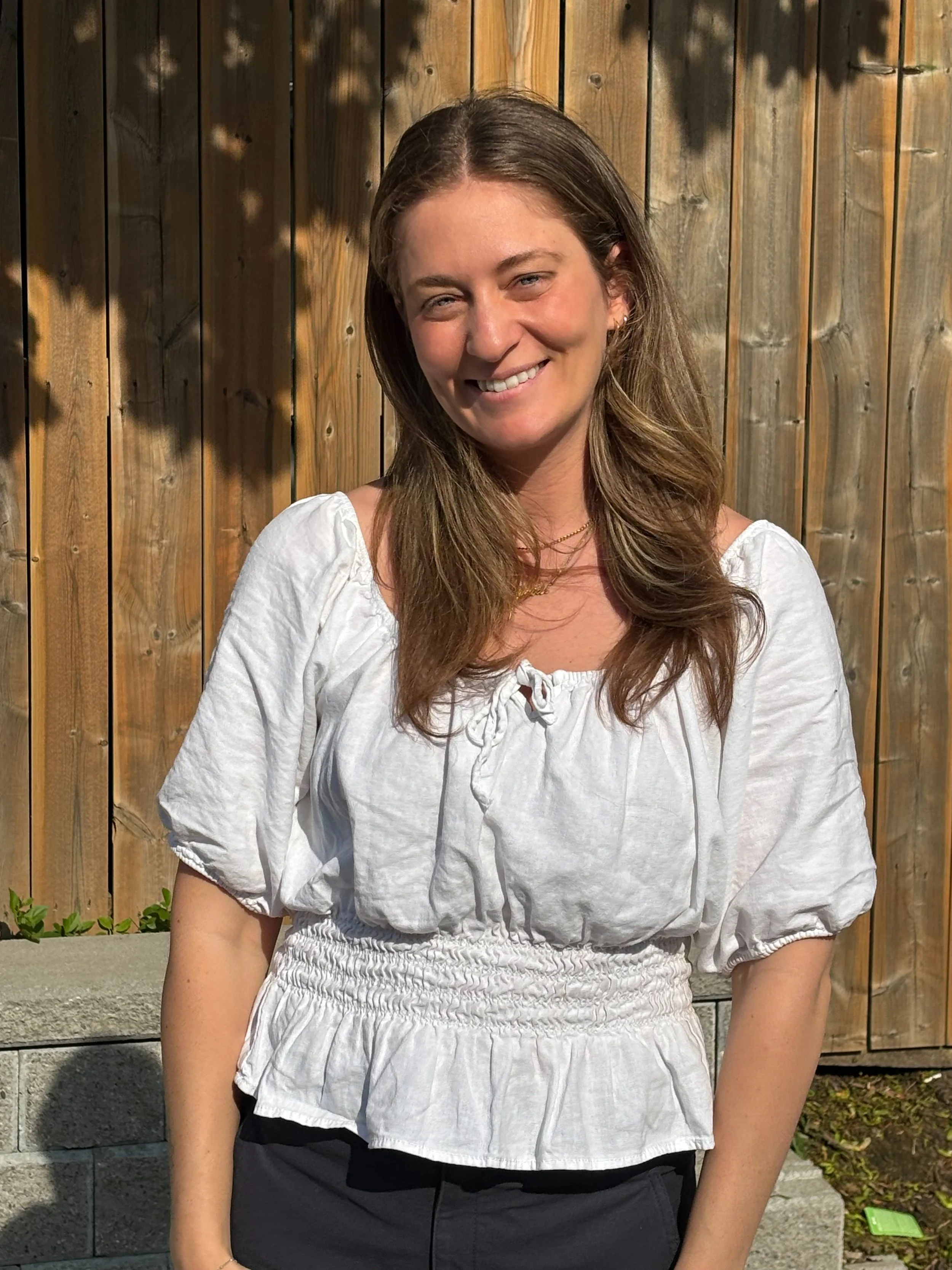 A smiling woman with long brown hair in a white ruched blouse standing outdoors in front of a wooden fence.