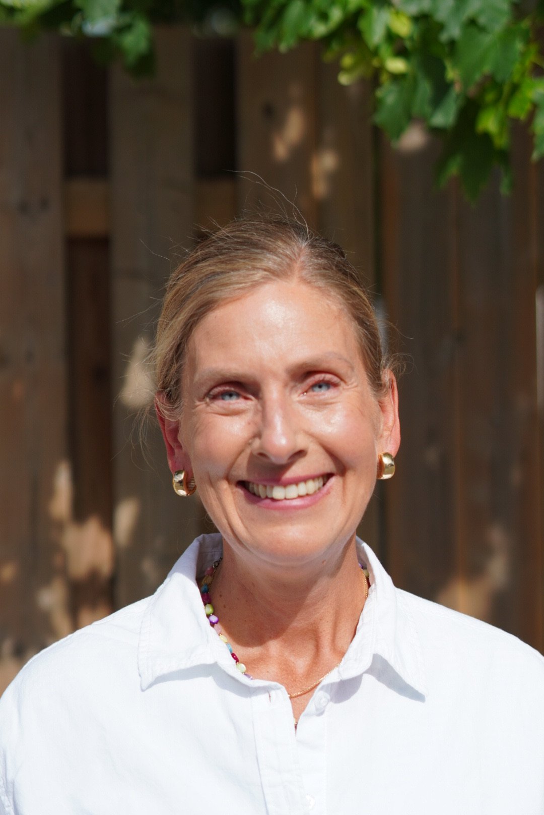 A woman smiling outdoors, wearing a white shirt, gold earrings, colorful necklace, and a gold chain, with a wooden fence and green foliage in the background.