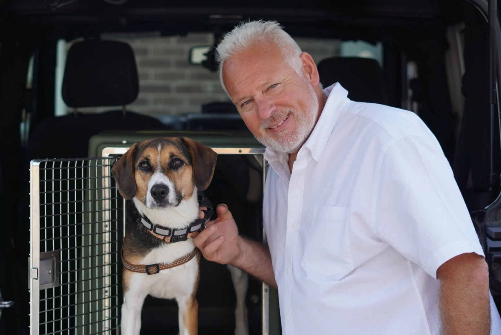 An older man with white hair and a white shirt smiling next to a beagle dog in a carrier, with the back of a vehicle in the background.