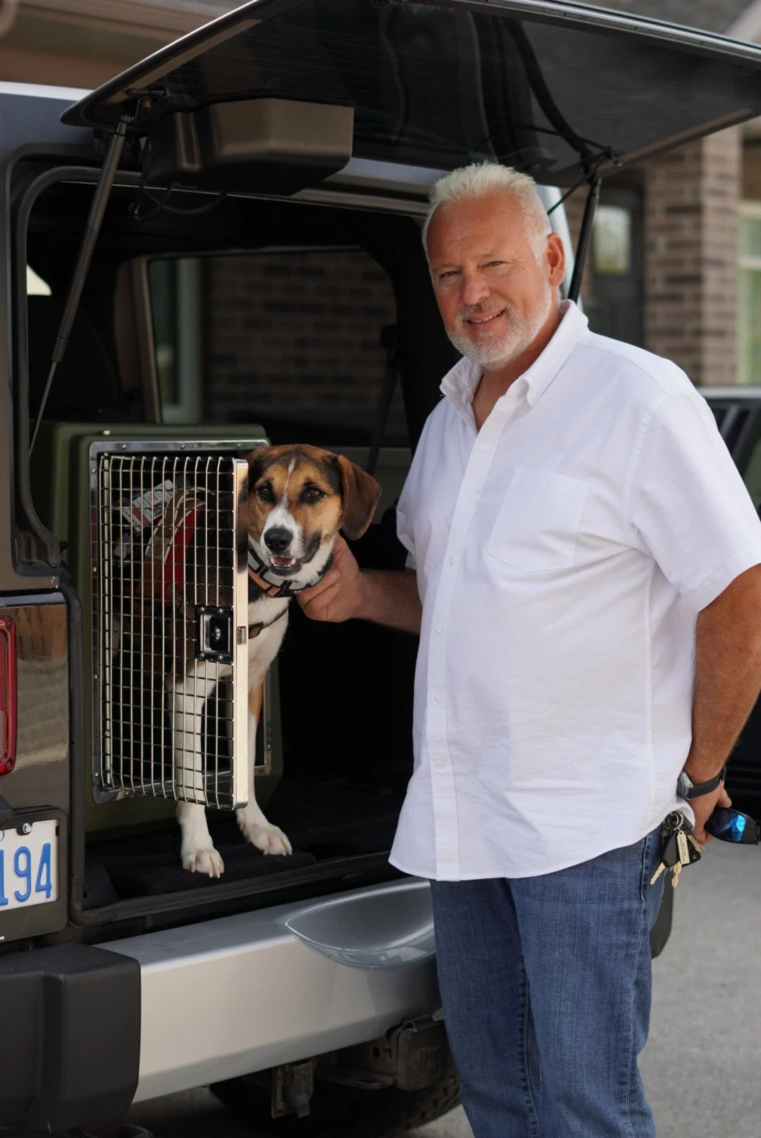 A man in a white shirt standing outside next to an open vehicle trunk with a dog in a crate, with a house in the background.