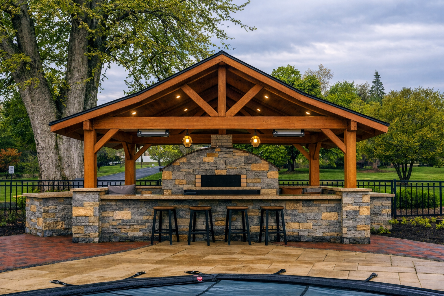 Outdoor pavilion with a stone fireplace, wooden roof structure, string lights, and bar stools, in a landscaped park setting.