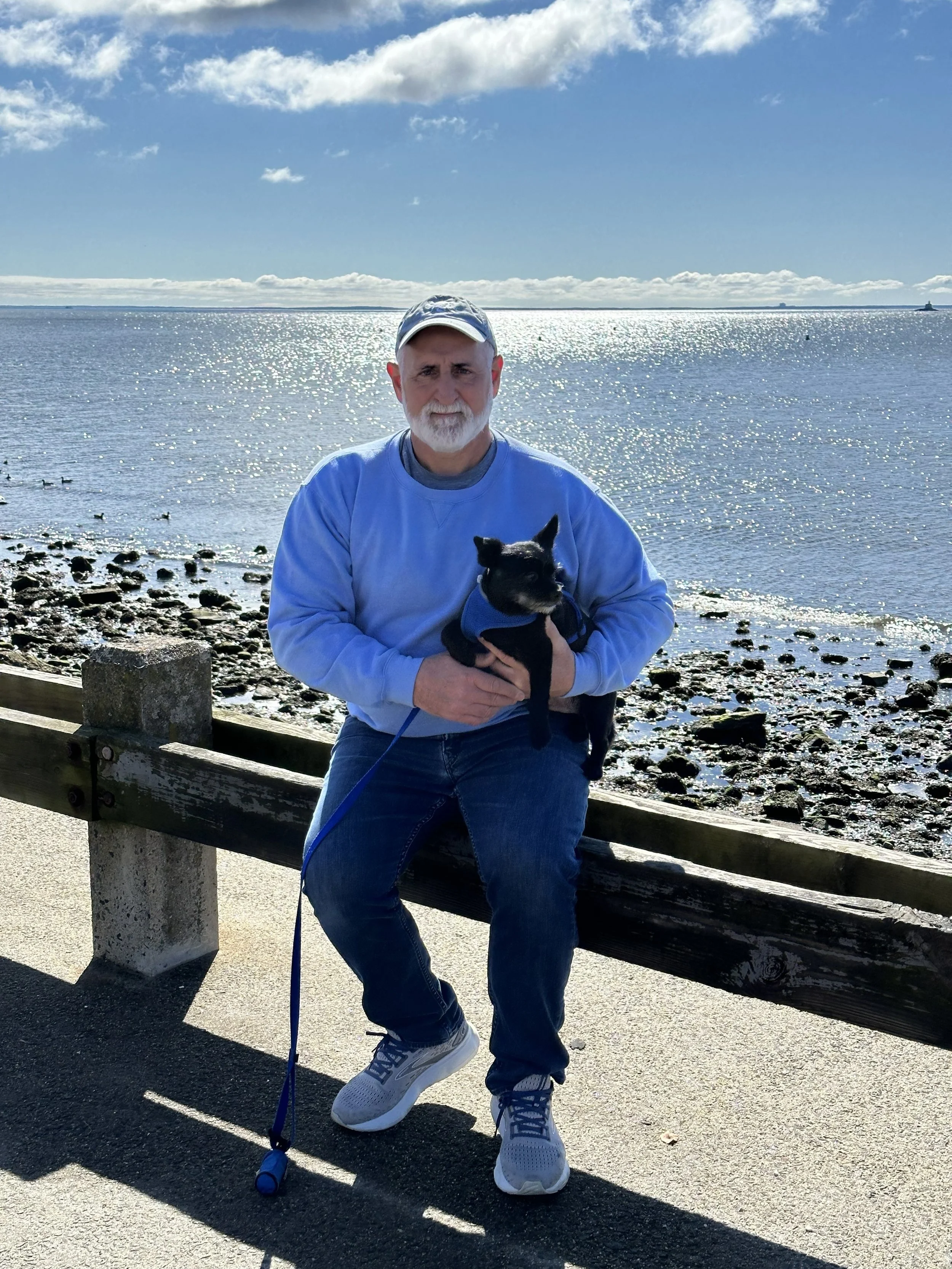A man with a white beard and mustache wearing a light blue sweatshirt, jeans, and sneakers, holding a small black dog, standing by a wooden railing on a walkway near a body of water under a partly cloudy sky.