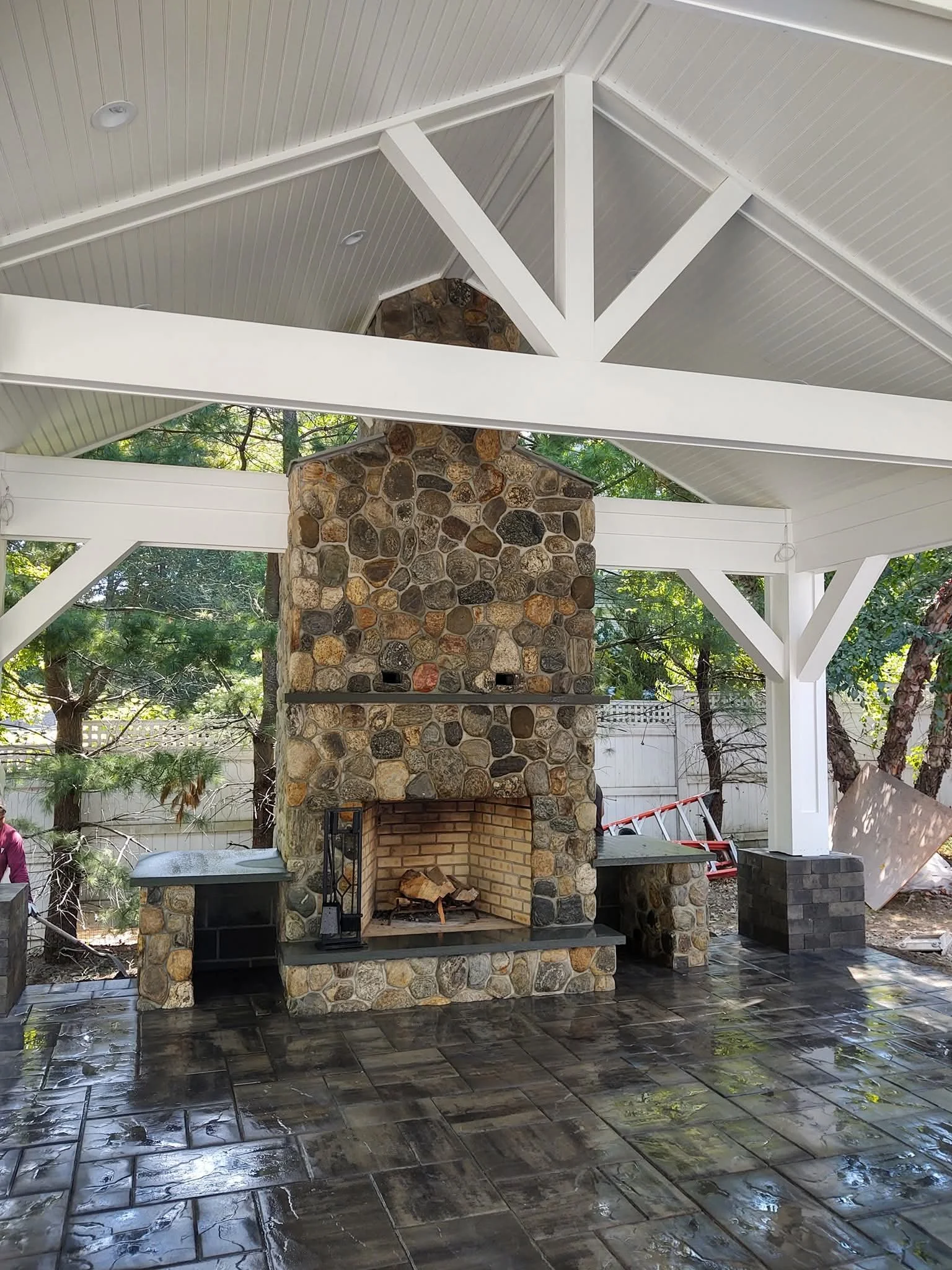 An outdoor patio area featuring a stone fireplace with a brick interior, under a white wooden pavilion with a vaulted ceiling. The floor is made of wet, shiny dark tiles, and there are trees and a white fence in the background.