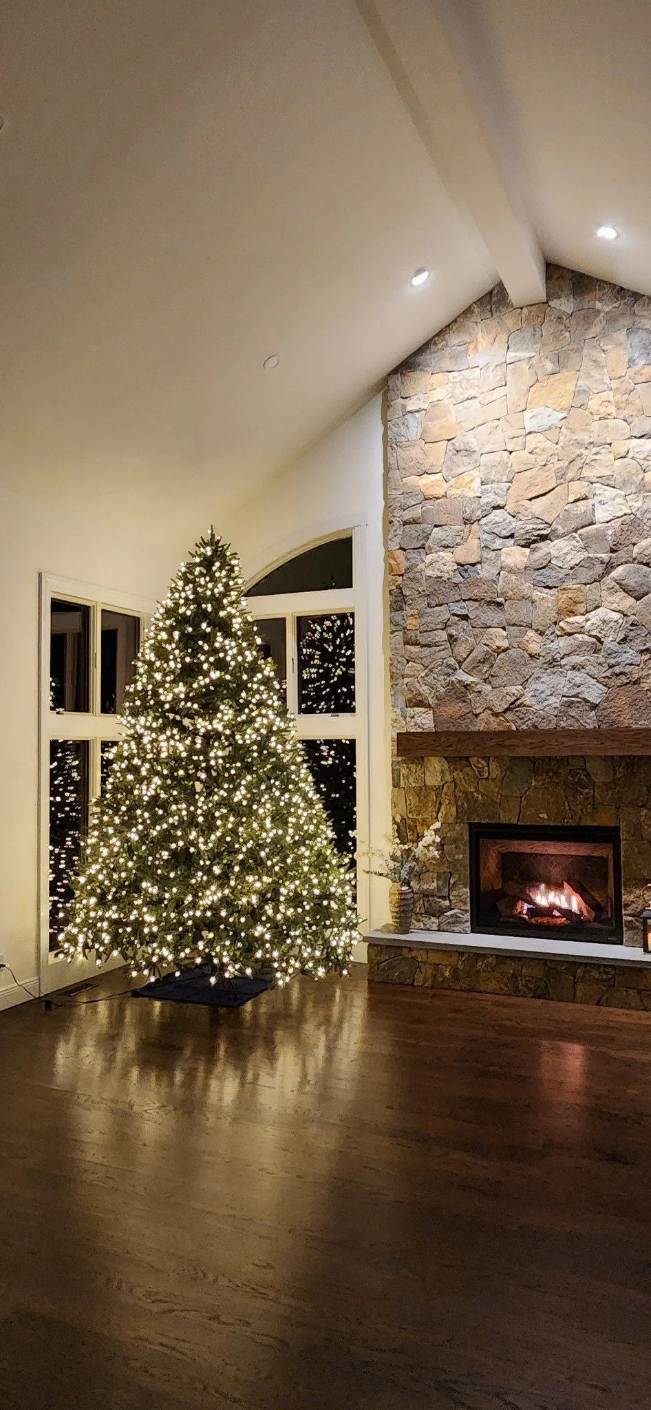Decorated Christmas tree with white lights in a living room with a stone fireplace, a vase with flowers, and a large arched window.