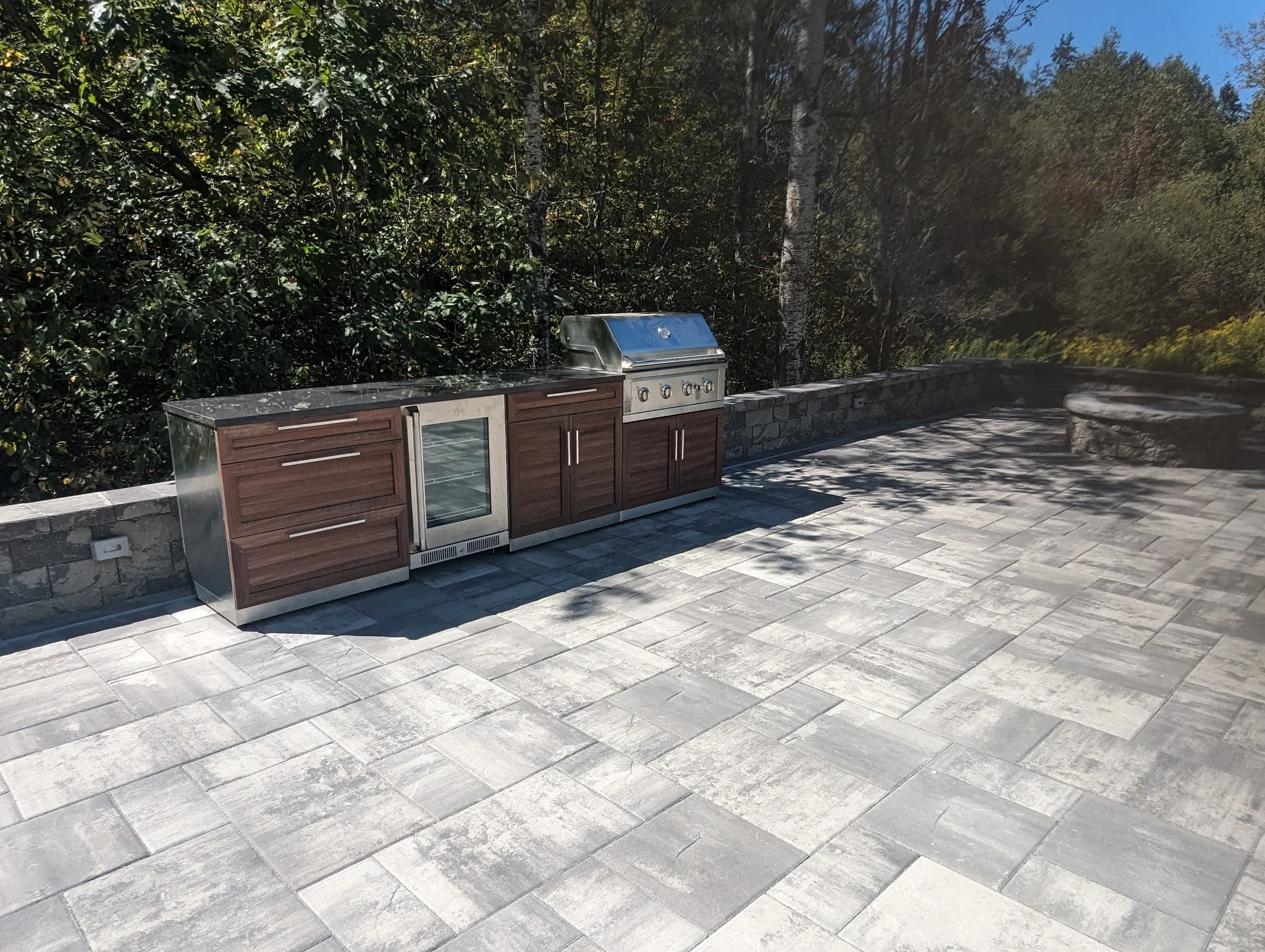 Outdoor patio with a built-in stainless steel grill, wooden cabinet doors, and a stone wall, with trees and blue sky in the background.