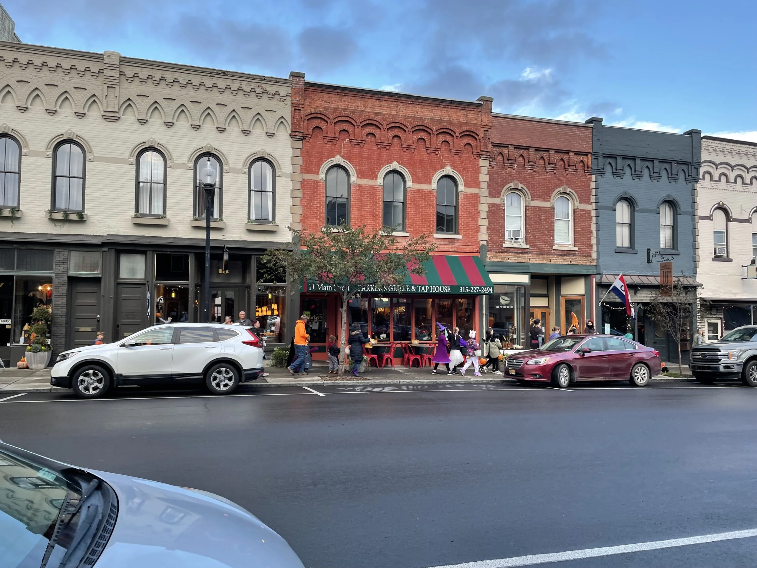 A row of colorful historic brick buildings with large arched windows on a city street. People are walking outside a restaurant with a green and red striped awning, while cars are parked along the street.