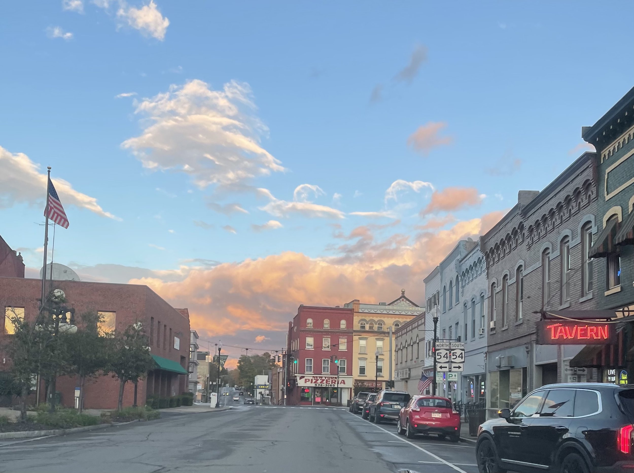 Street view with buildings, cars parked on the side, a neon sign that reads 'TAVERN', a pizza restaurant sign, and an American flag flying, under a sky with clouds at sunset.
