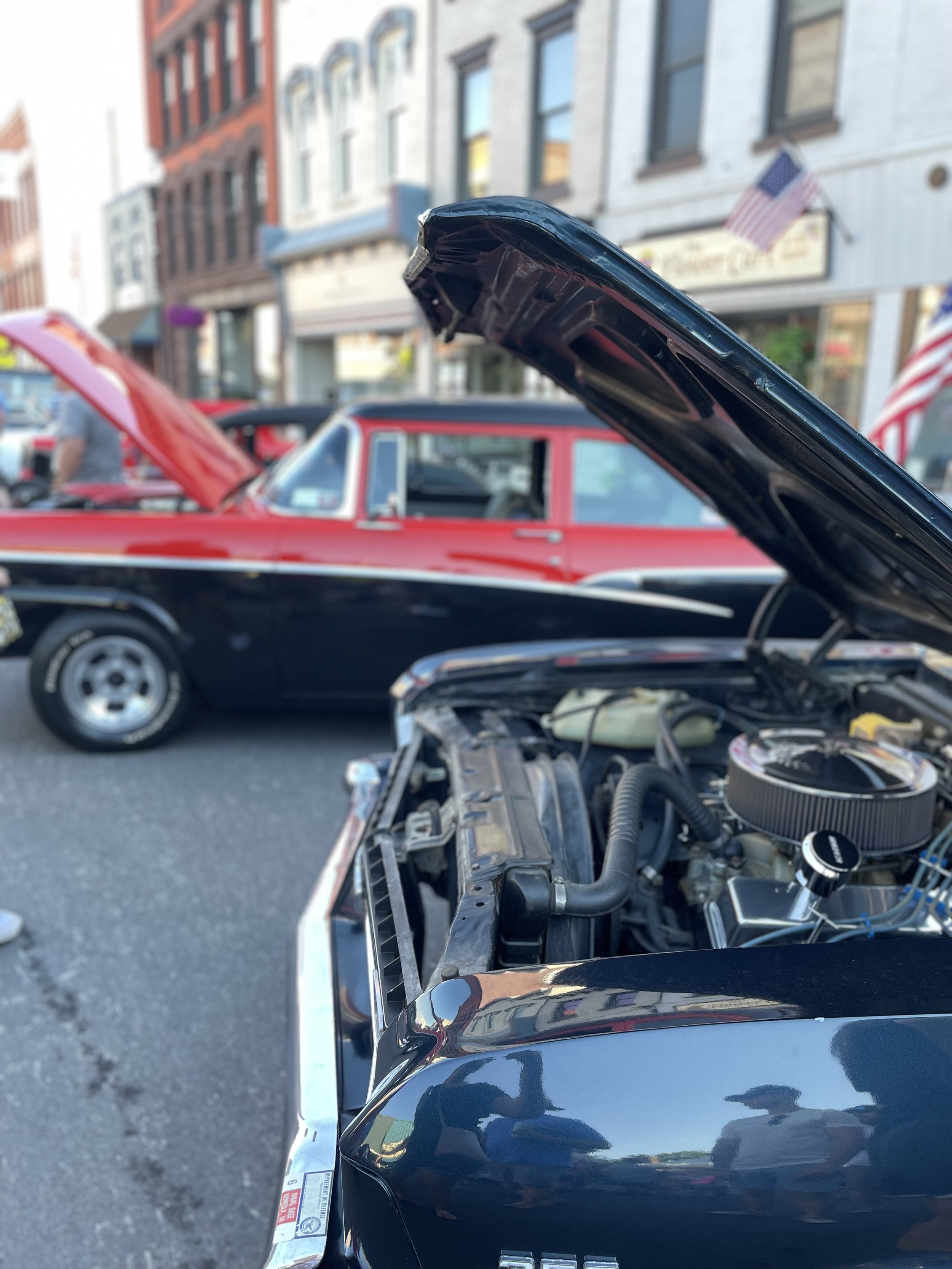 Close-up of an open car hood showing the engine compartment, with a classic red car parked parallel in the background on a street lined with colorful buildings.