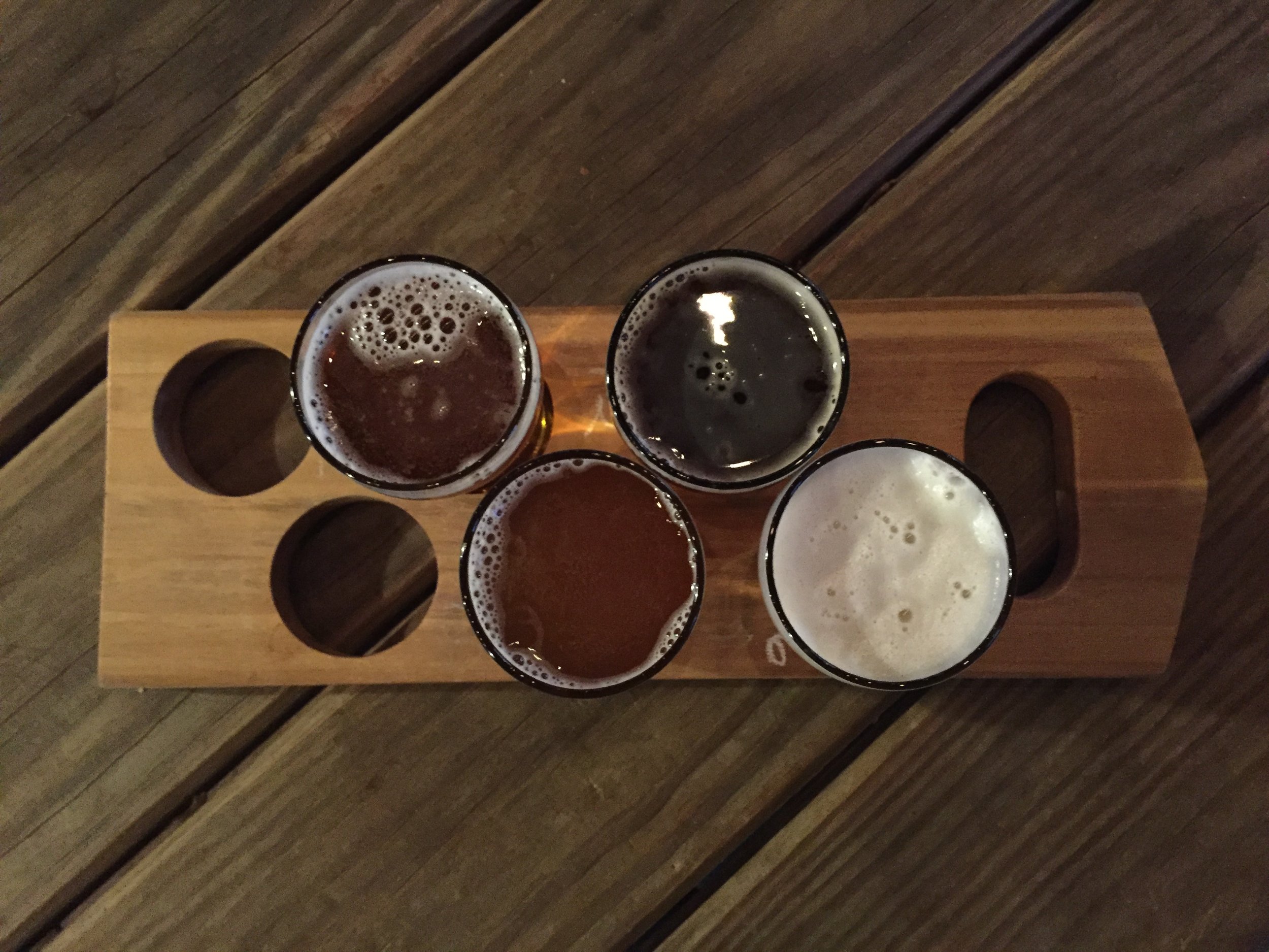 Top view of a wooden flight flight tray with five glasses of beer on it. The tray is on a wooden table or floor.