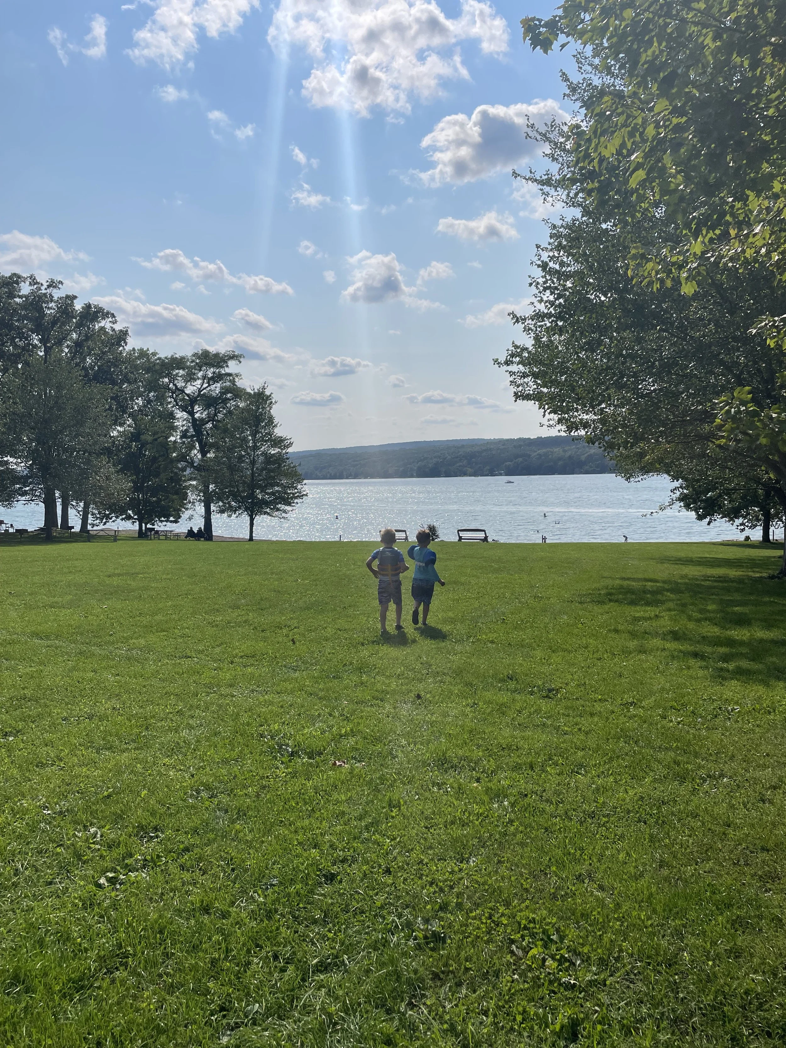 Two children stand on a lush green lawn near a large body of water, with trees on either side and a partly cloudy sky overhead.