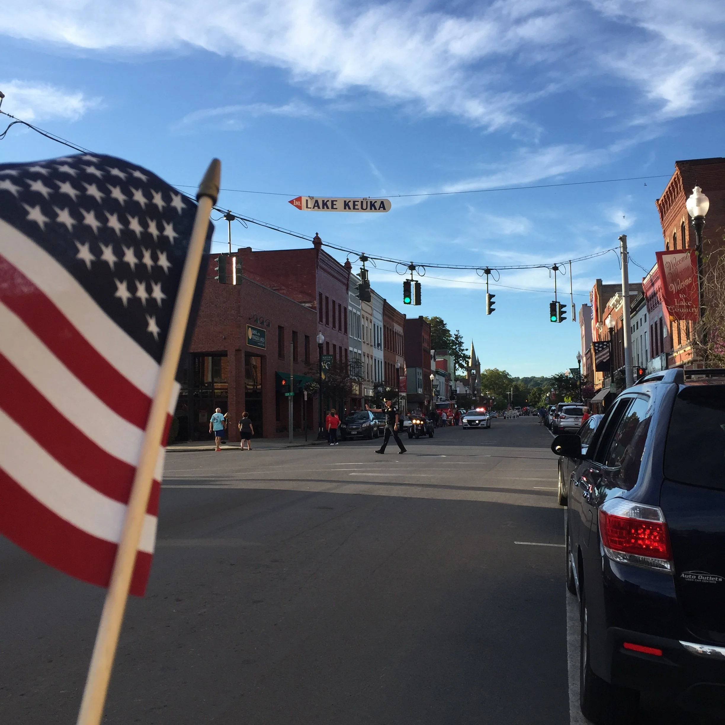 Street scene in Lake Keuka with buildings, people walking, cars parked and traffic lights, with a blue sky and some clouds above.