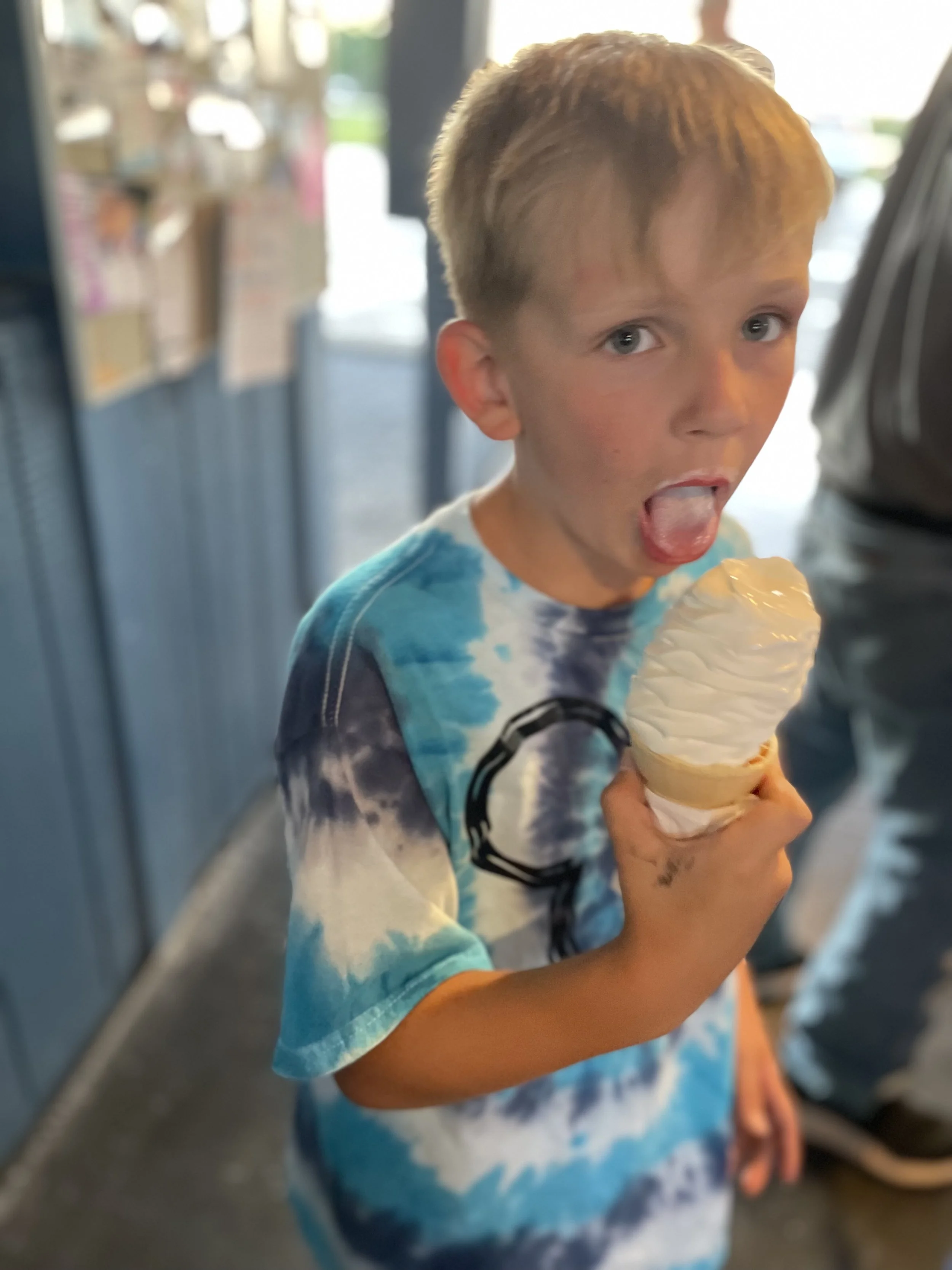A young boy with short light brown hair and blue eyes, wearing a tie-dye shirt, is licking a vanilla ice cream cone. The background shows a blurry cafeteria or shop with people and shelves.