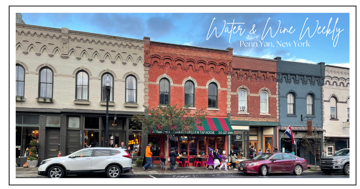 Street scene in Penn Yan, New York showcasing historic brick buildings, a restaurant with red and green striped awning, people walking on the sidewalk, parked cars, and a blue sky with some clouds.