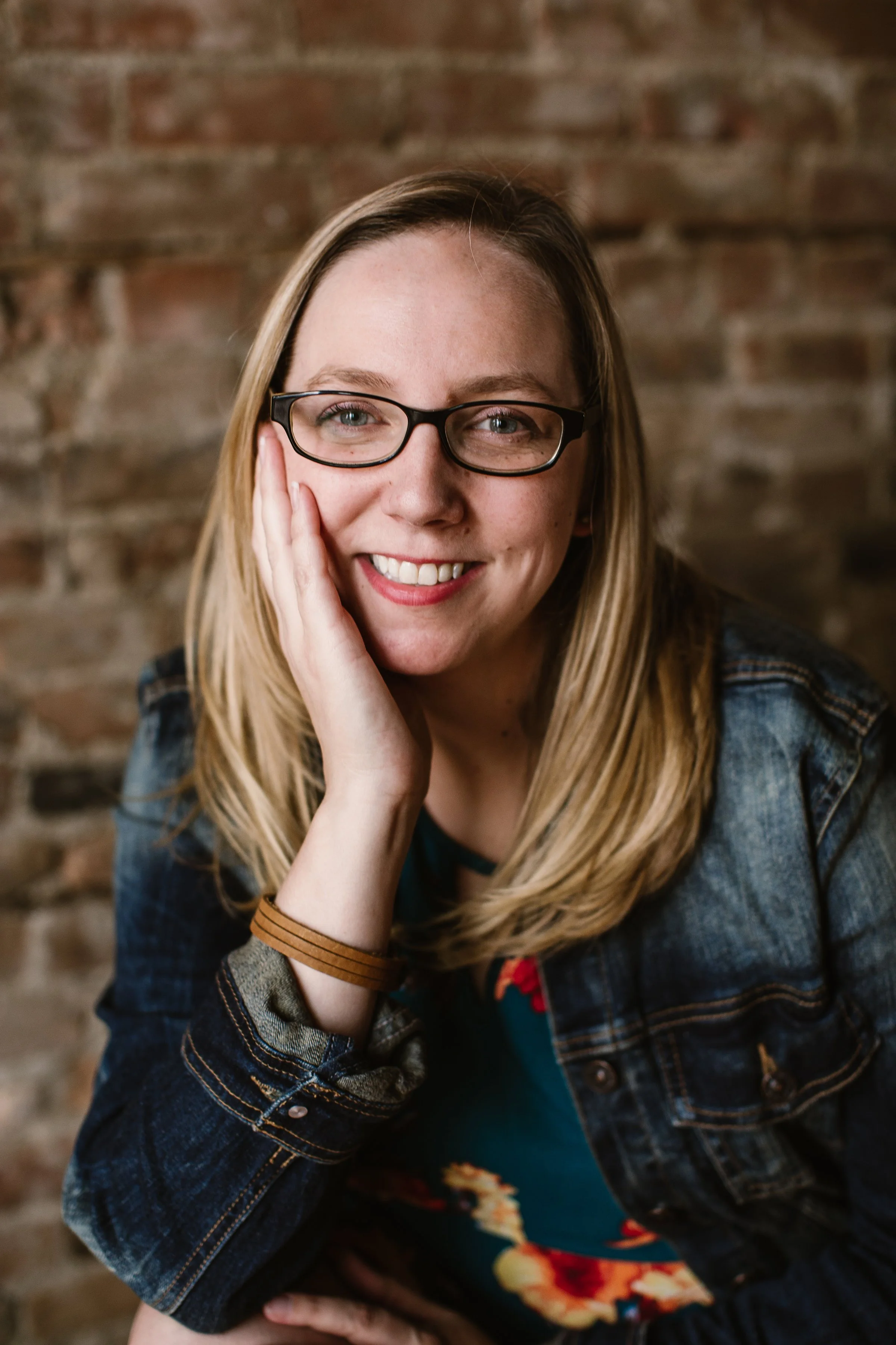 A woman with blonde hair and glasses smiling, resting her face on her hand, against a brick wall background.