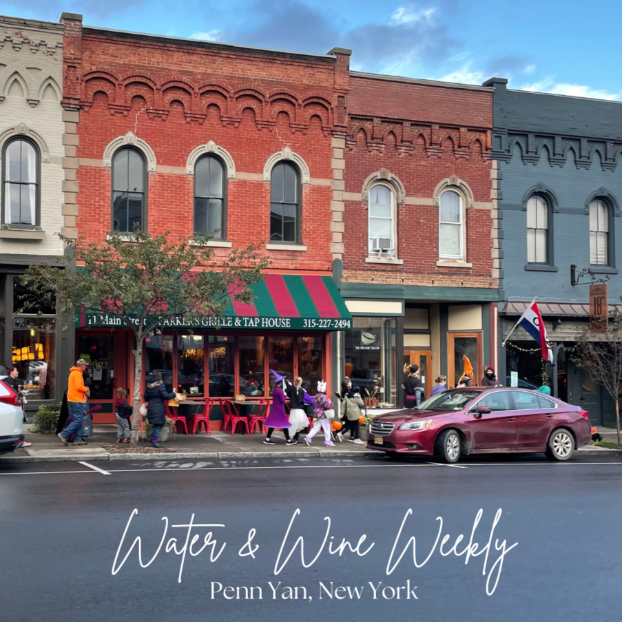 Street scene in Penn Yan, New York, featuring a row of brick buildings with storefronts, including a tap house with a green and red striped awning, pedestrians in Halloween costumes, parked cars, and a cloudy sky.