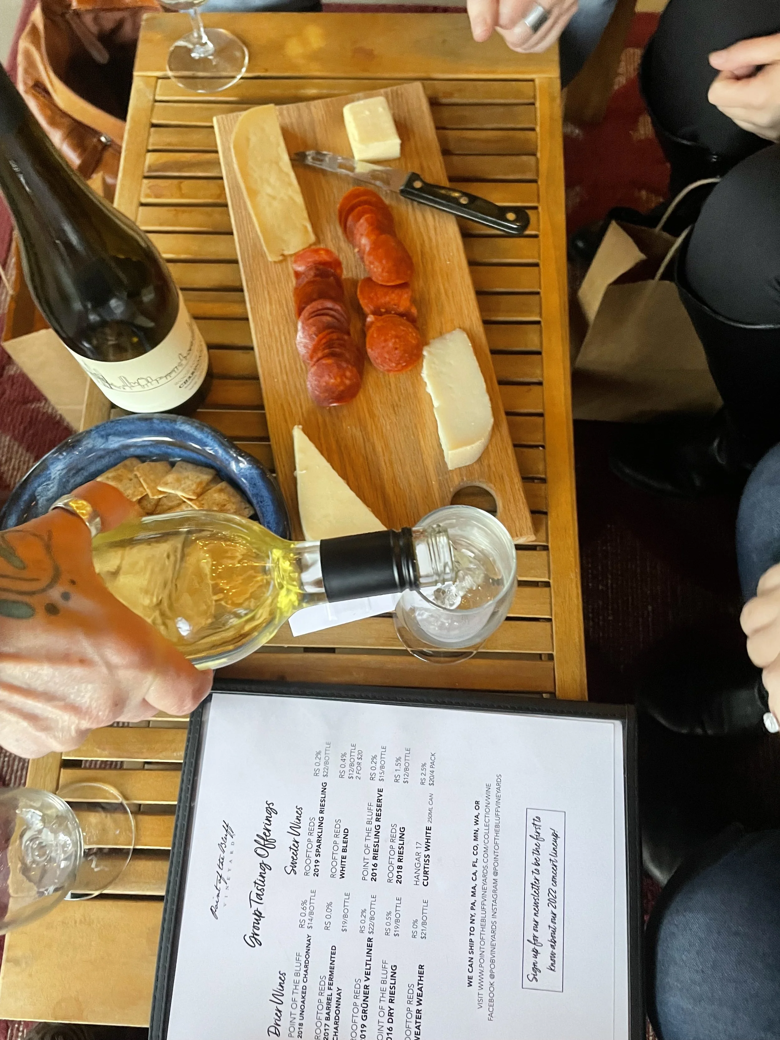 A person pouring a glass of white wine at a table with a cheese and charcuterie board. The board contains slices of cheese and salami. There is a bottle of wine, a menu, and other glasses on the table.