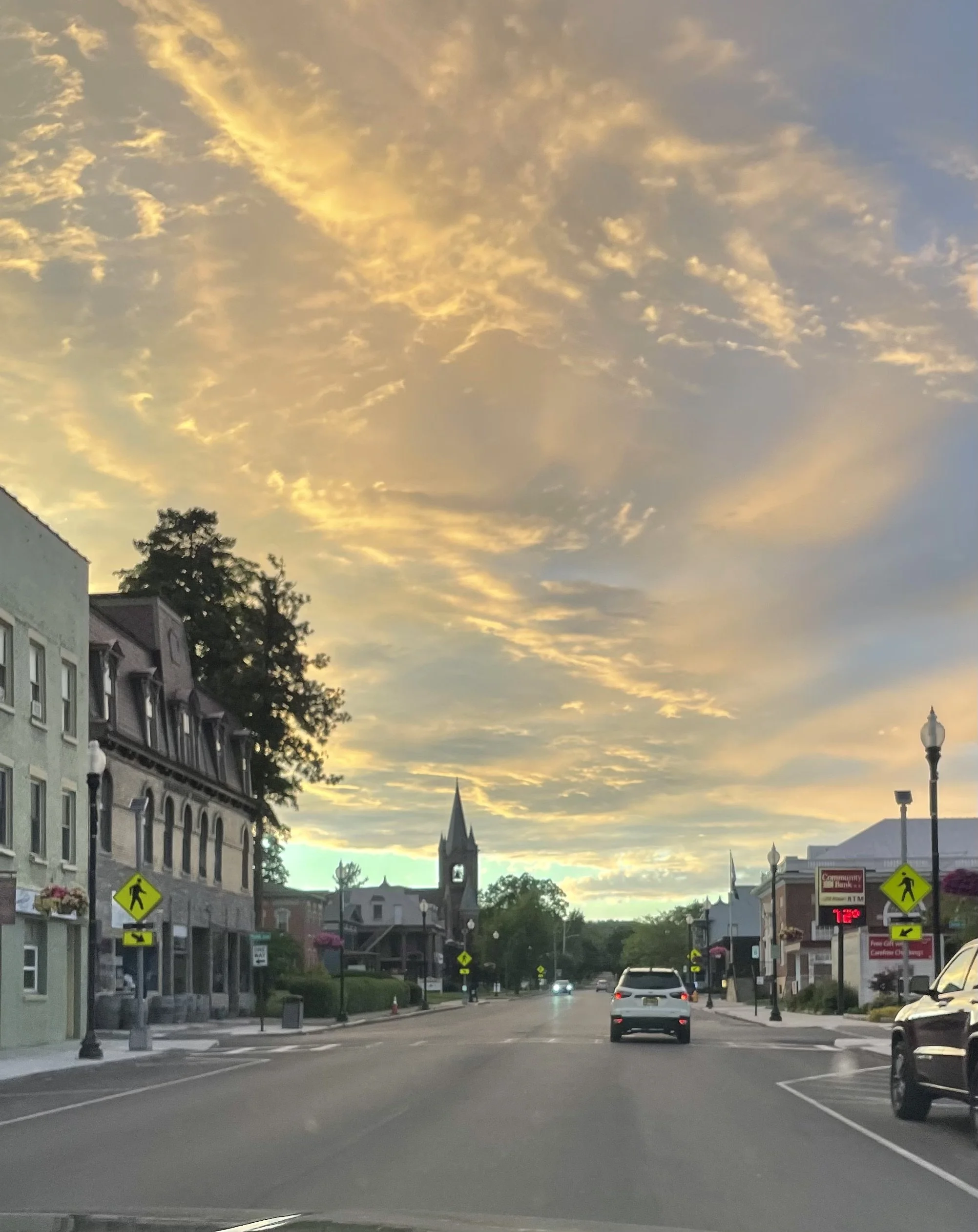 A street view during sunset with buildings on the left and cars on the road. There are yellow pedestrian crossing signs and a digital sign displaying temperature 72°F and other information. The sky has clouds illuminated by the setting sun.