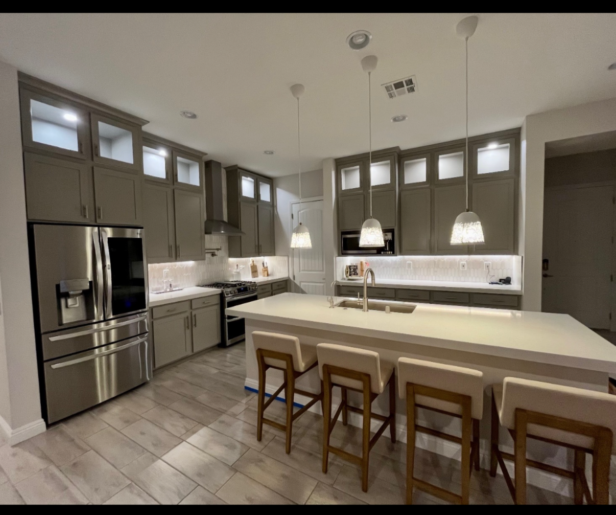 Modern kitchen with gray cabinetry, stainless steel refrigerator, built-in microwave, gas stove, white countertops, island with four barstools, pendant lighting, and light-colored tiled floors.